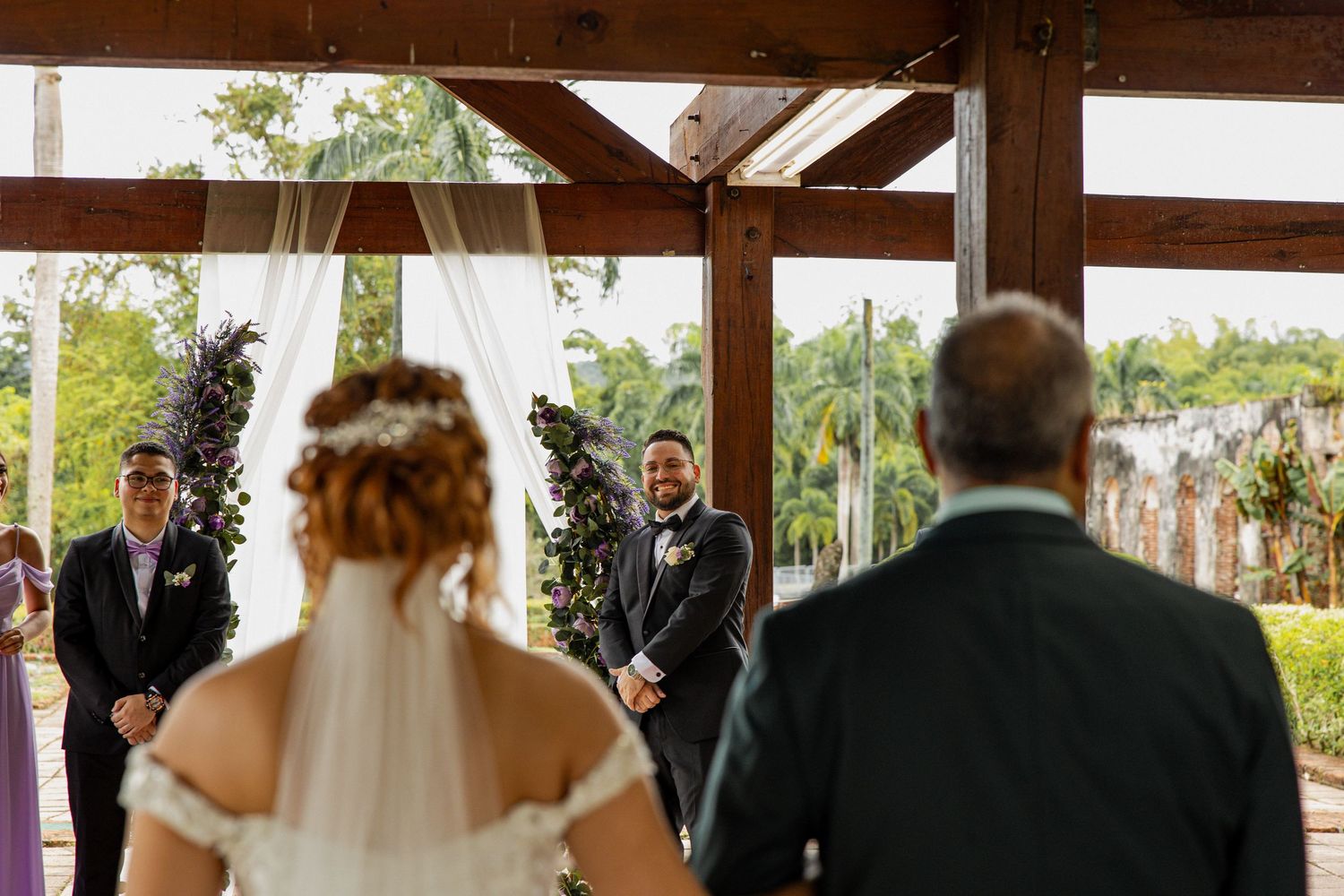 Bride in white gown and veil walks toward groom at outdoor wedding ceremony under rustic wooden pavilion.