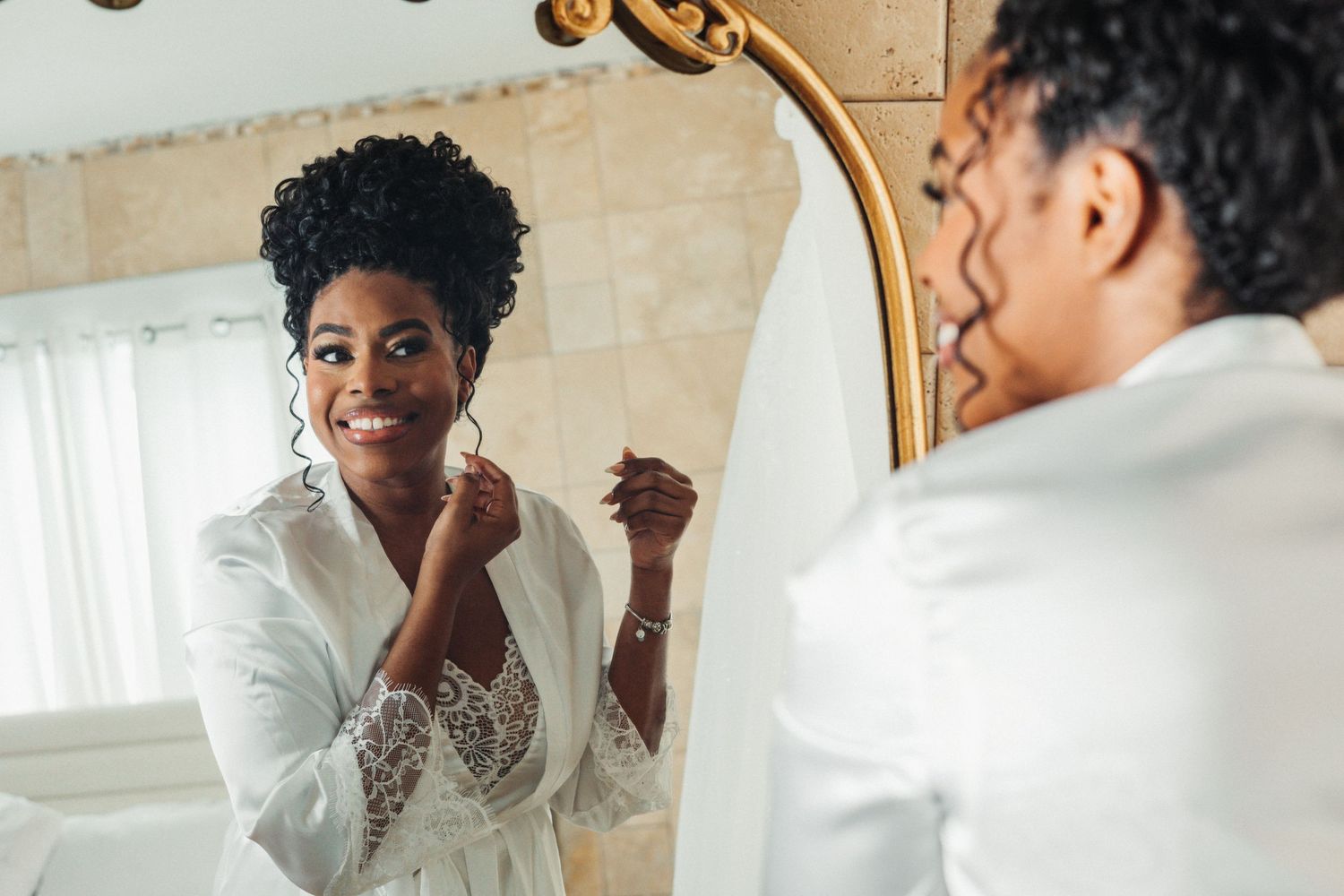 A smiling Black woman in a white lace robe puts on earrings while looking at her reflection in an ornate mirror.