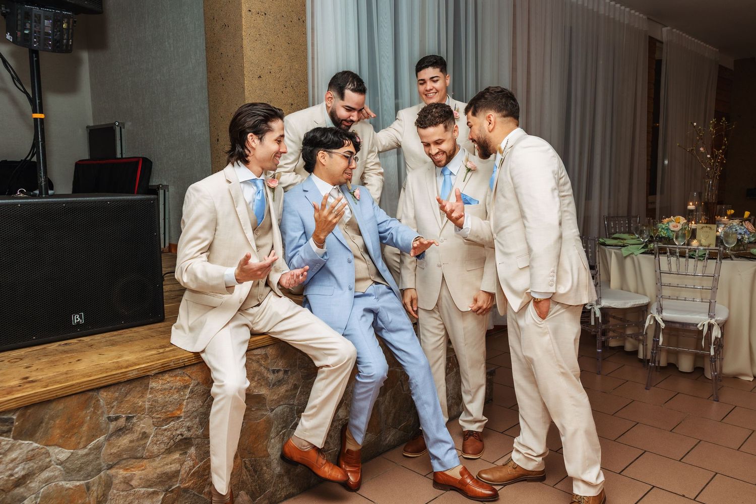 Groom in light blue suit laughing with groomsmen in cream suits at wedding reception venue.