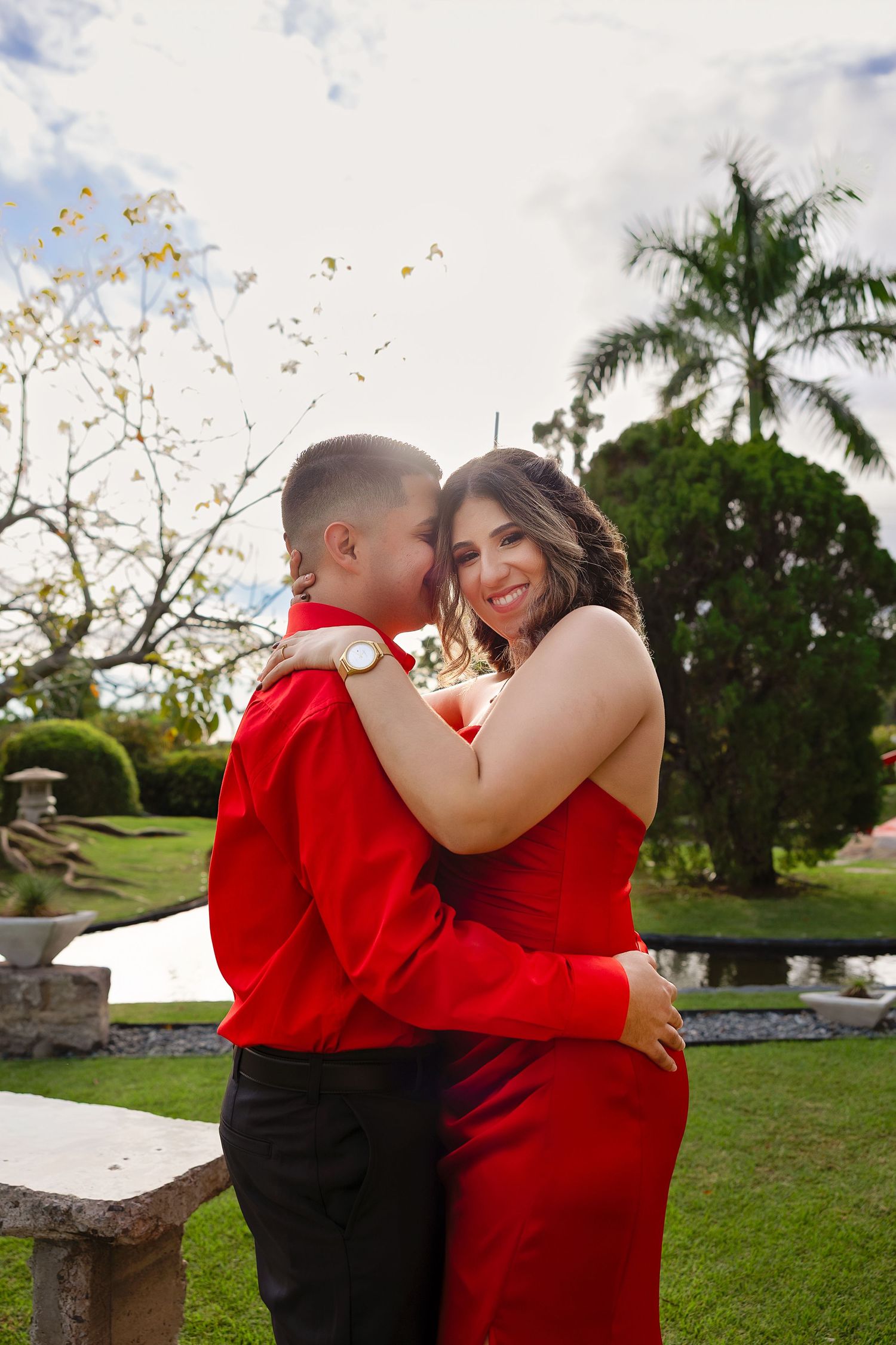 A couple in matching red outfits share an intimate moment in a tropical garden setting with palm trees.