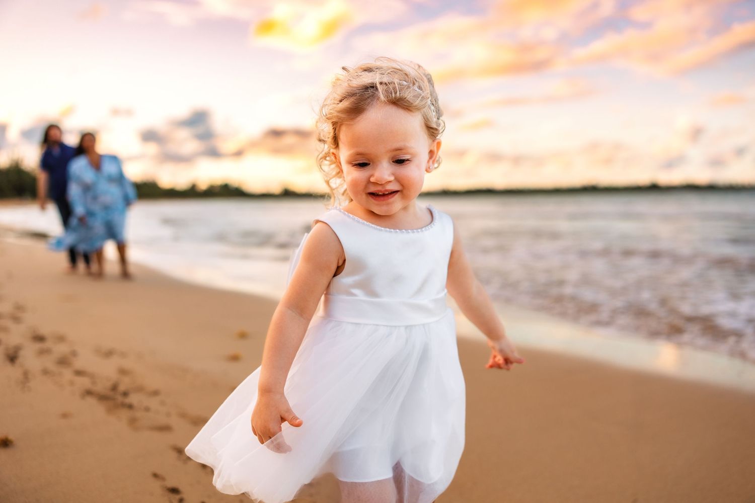 Joyful toddler girl in white dress running on sandy beach at sunset with family in background.
