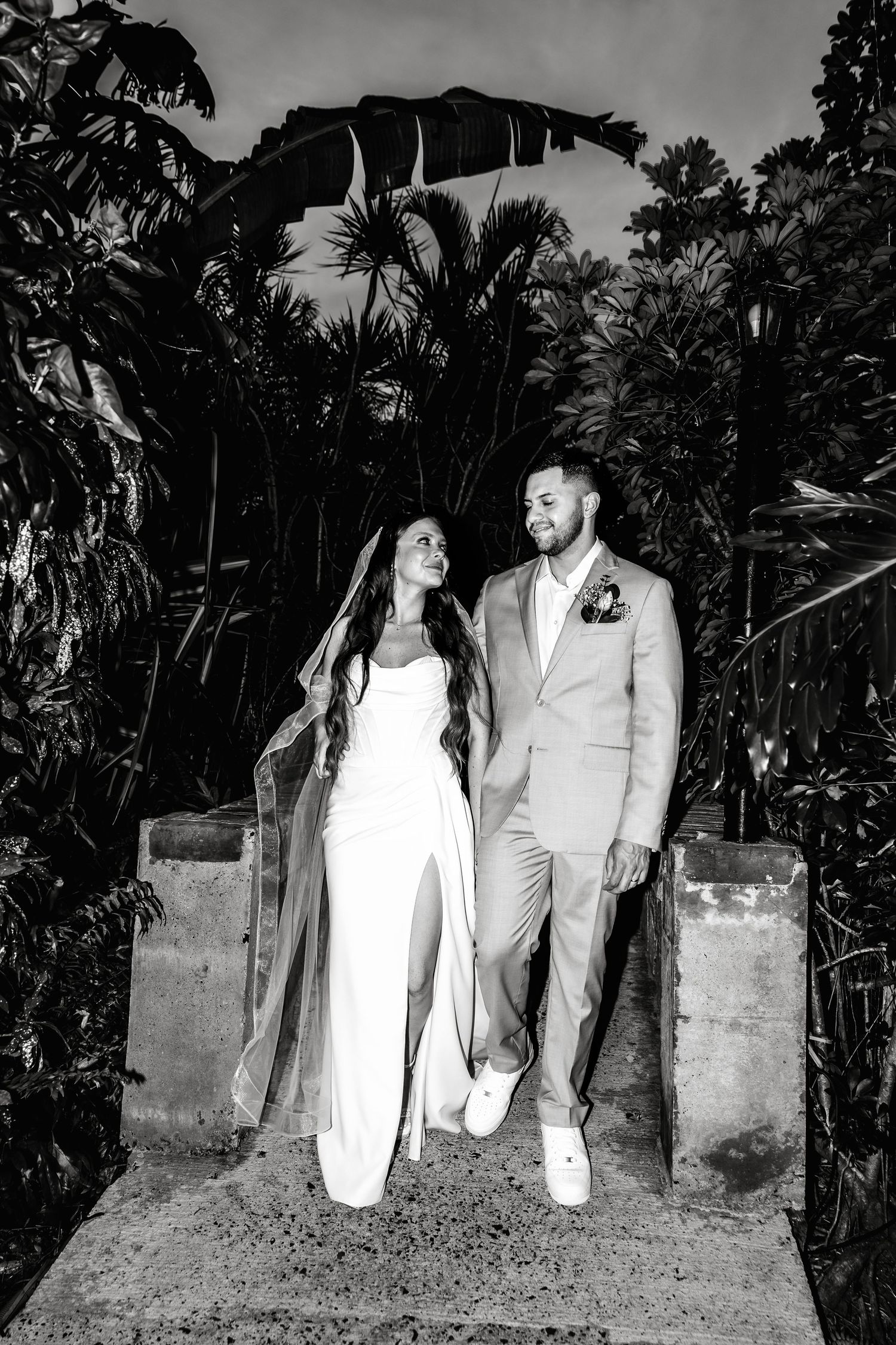 Black and white photo of a bride and groom walking together outdoors surrounded by tropical plants at night.