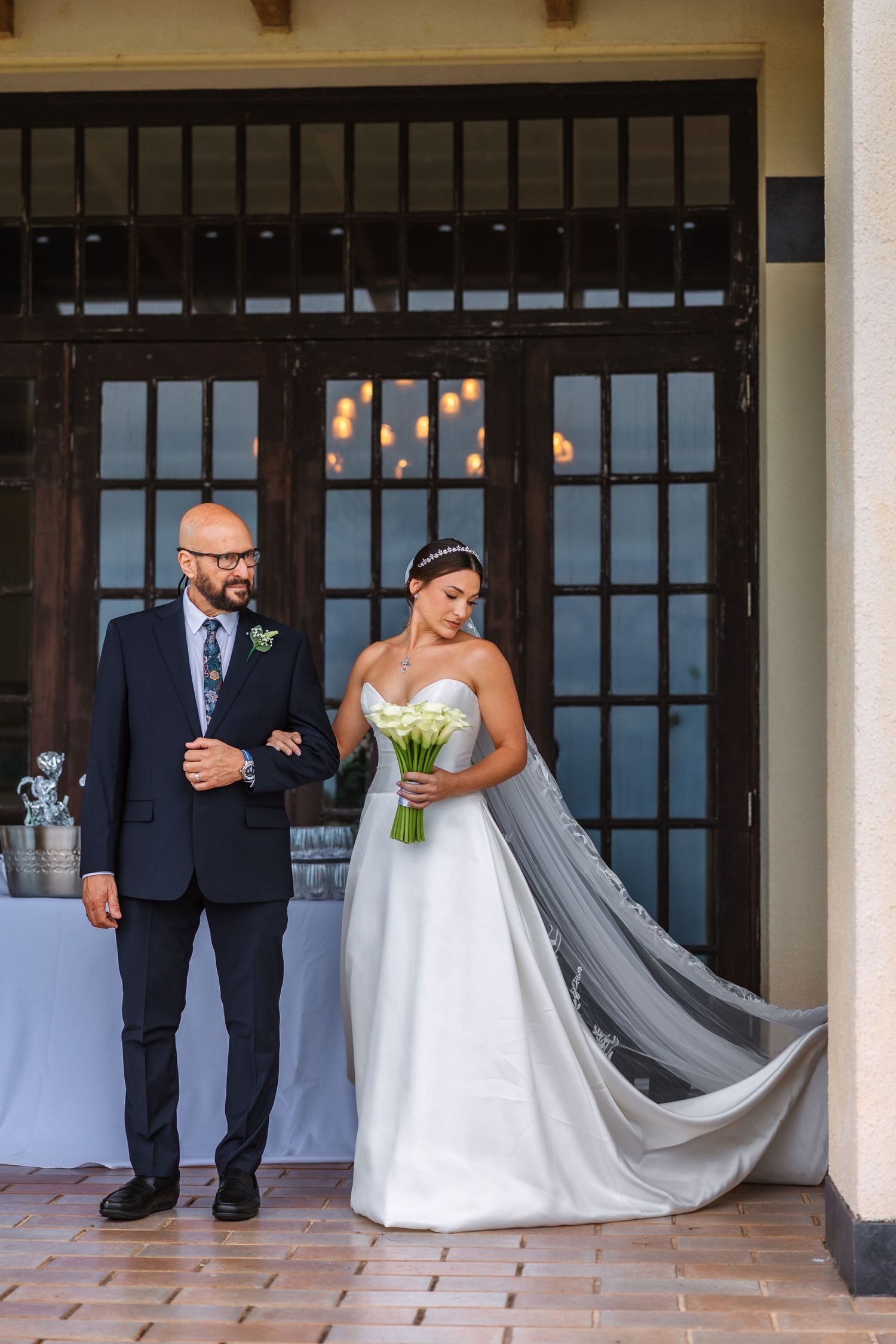 Bride in white strapless gown holding bouquet stands with suited man before elegant French doors.