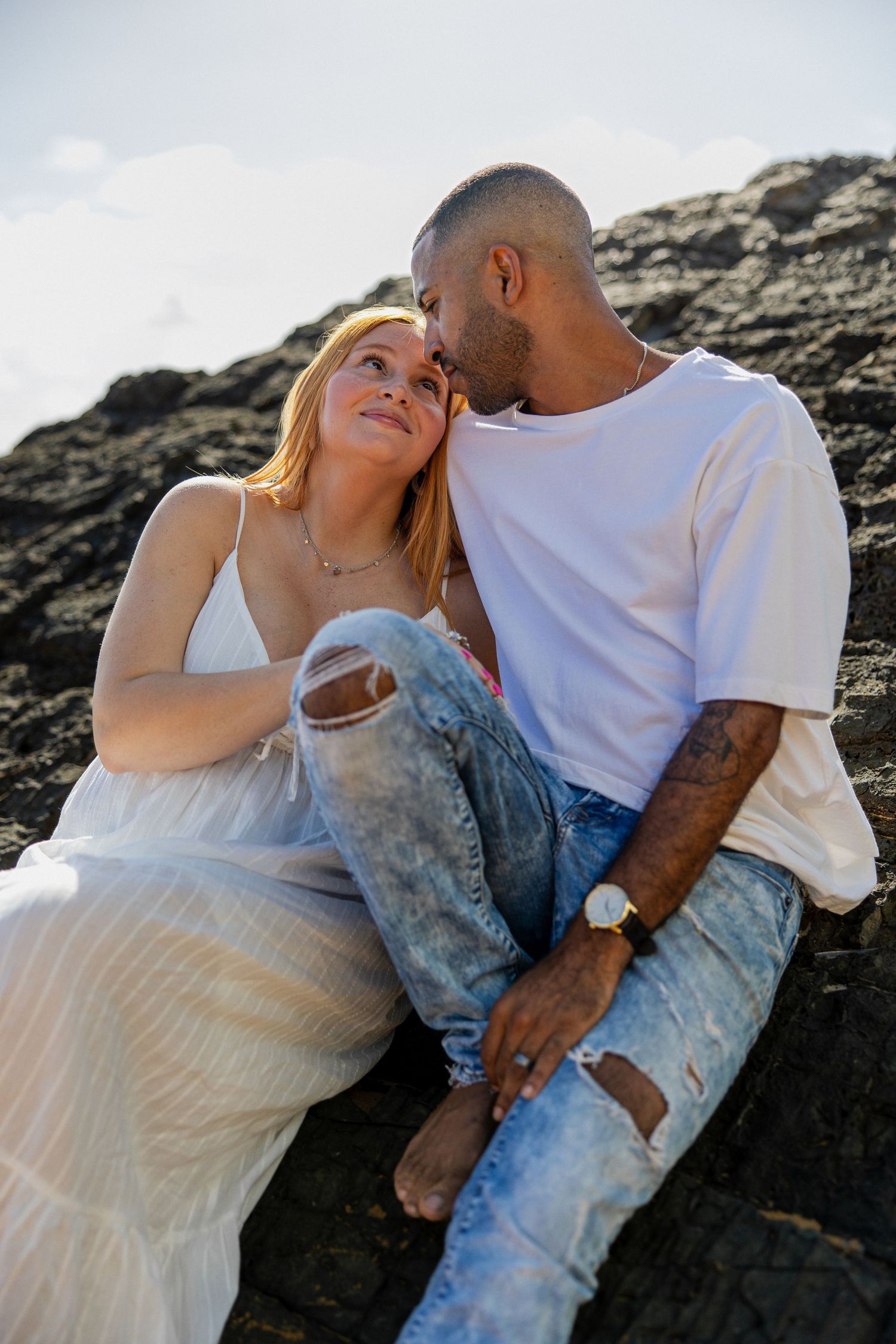 A couple sits together on rocks, sharing a tender moment, woman in white dress and man in white tee and ripped jeans.