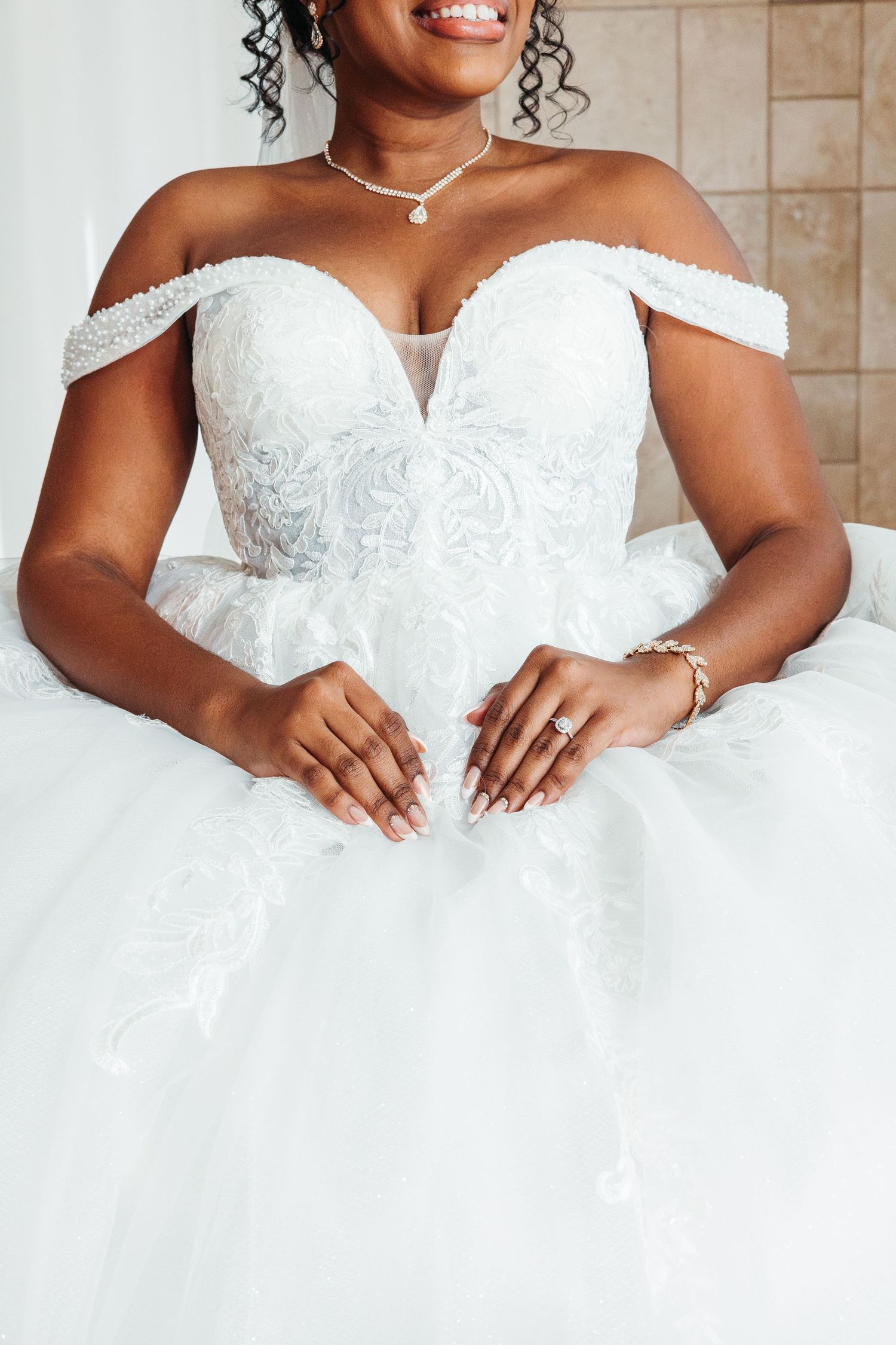 Black bride in off-shoulder white lace ballgown wedding dress with pearl necklace and diamond ring, smiling.