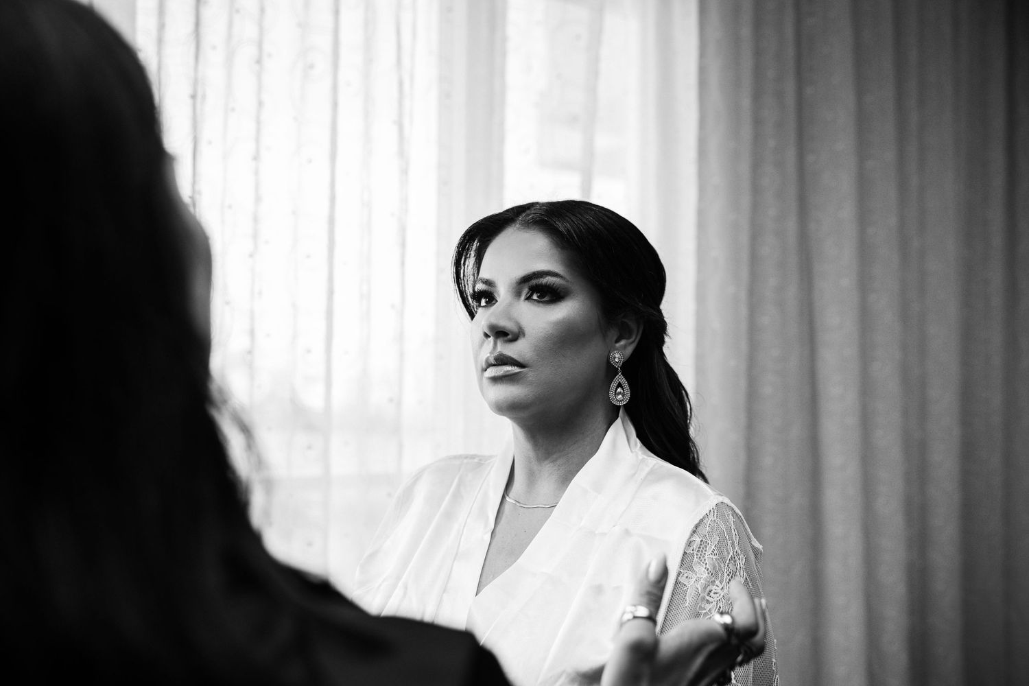 Black and white photo of a bride in a white robe getting ready, wearing elegant earrings near a window.