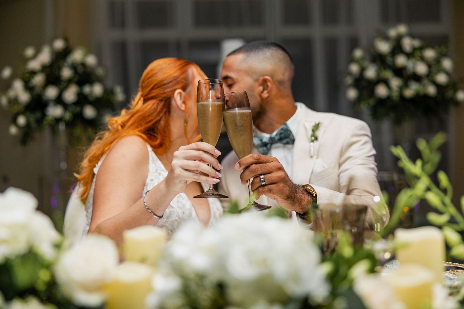 Bride and groom share a romantic kiss while toasting with champagne flutes at their elegant wedding reception.