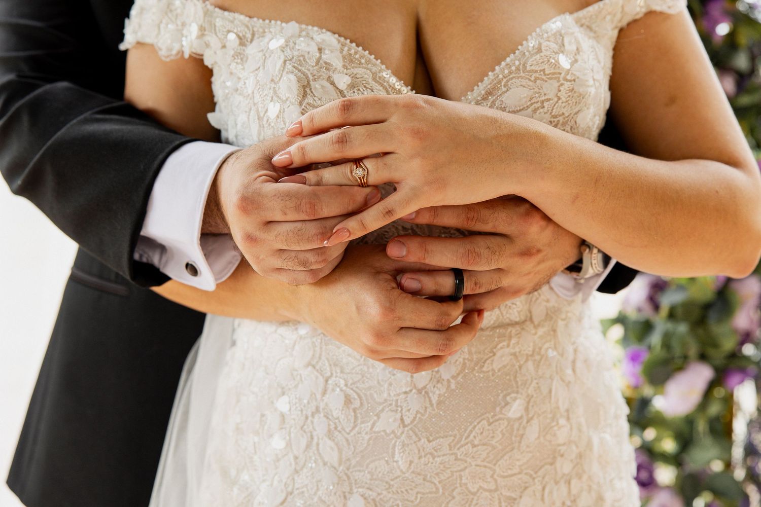 Bride and groom showing wedding rings, hands intertwined over lace bridal gown.