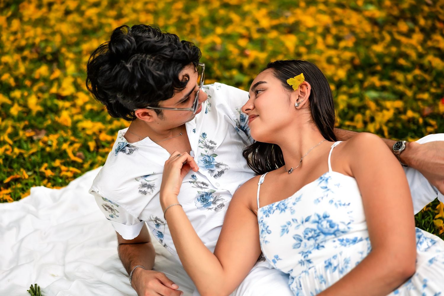 Couple in floral outfits lying on white blanket surrounded by yellow flowers, gazing at each other romantically.