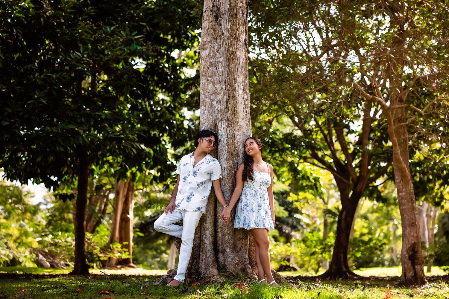 A couple poses playfully near trees in a park during a sunny outdoor photoshoot.