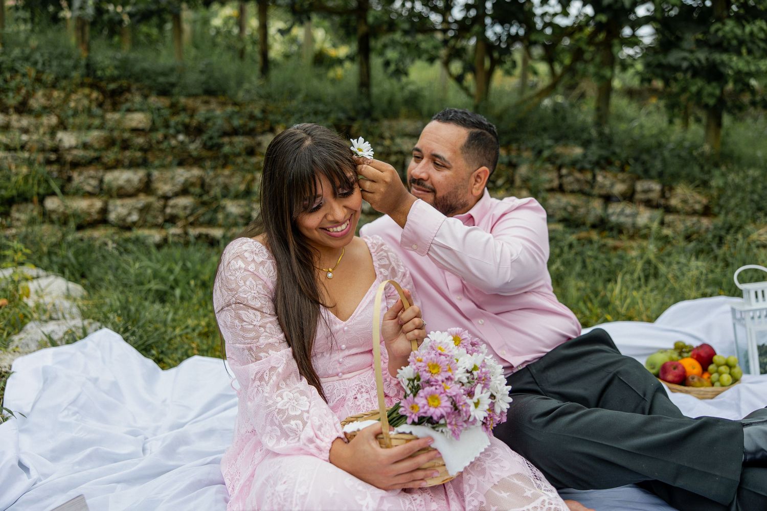 Couple sharing a romantic outdoor picnic, man placing flower in woman's hair, pink dress, bouquet, fruit basket.
