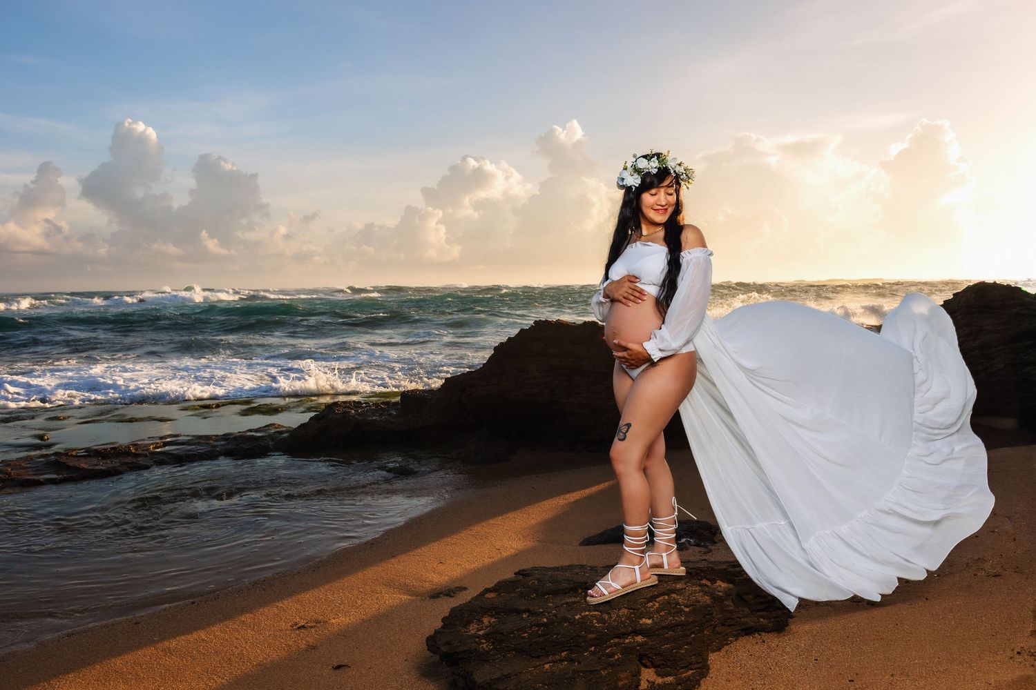 Pregnant woman in white flowing dress and floral crown poses on rocky beach at sunset with ocean waves behind her.