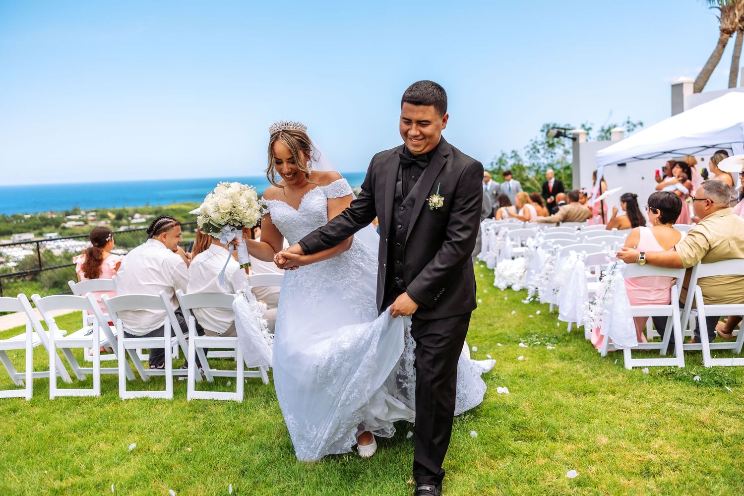 Bride and groom walk down outdoor wedding aisle on green lawn with ocean view and white chairs.