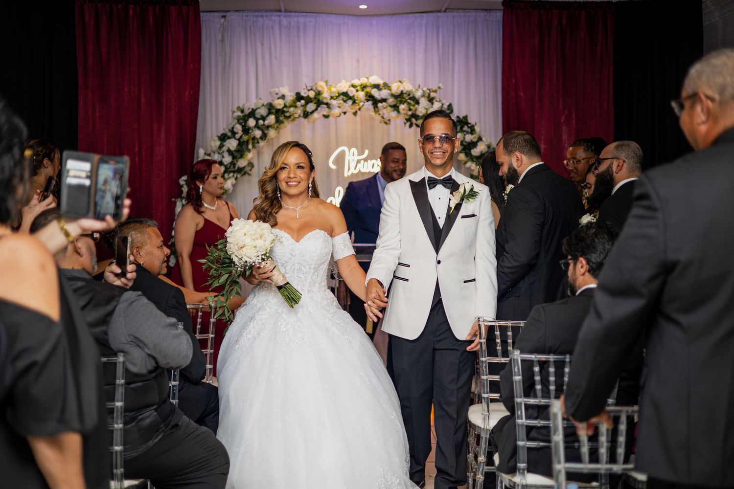 Newlyweds walking down the aisle under a floral arch with guests looking on.