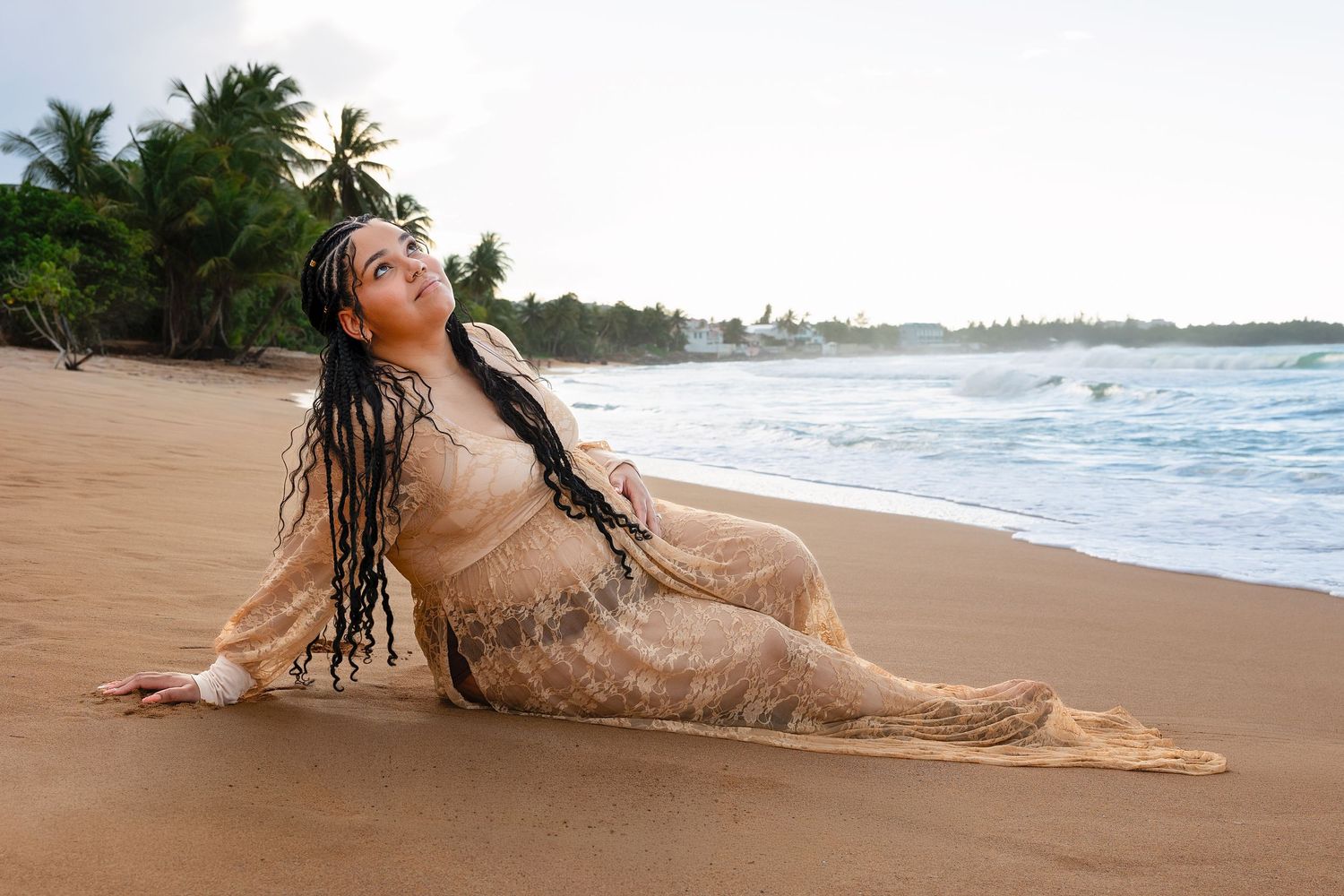 Woman in sheer beige dress with long braids poses gracefully on a tropical sandy beach by the ocean.