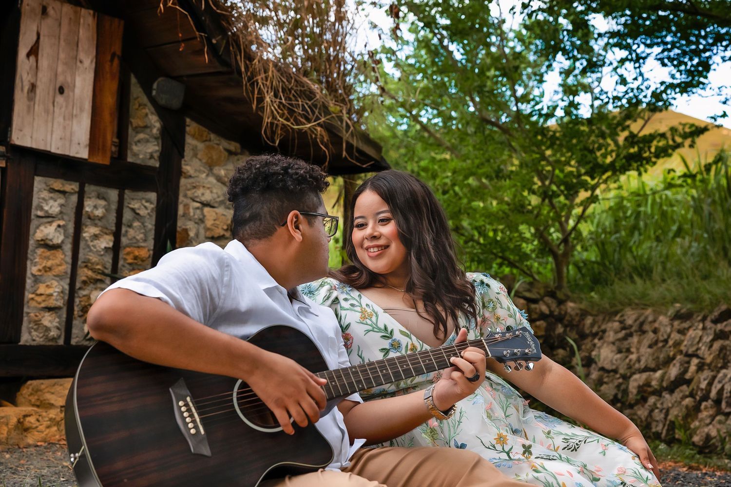 A young man plays acoustic guitar for a smiling woman outdoors near a rustic stone building surrounded by greenery.
