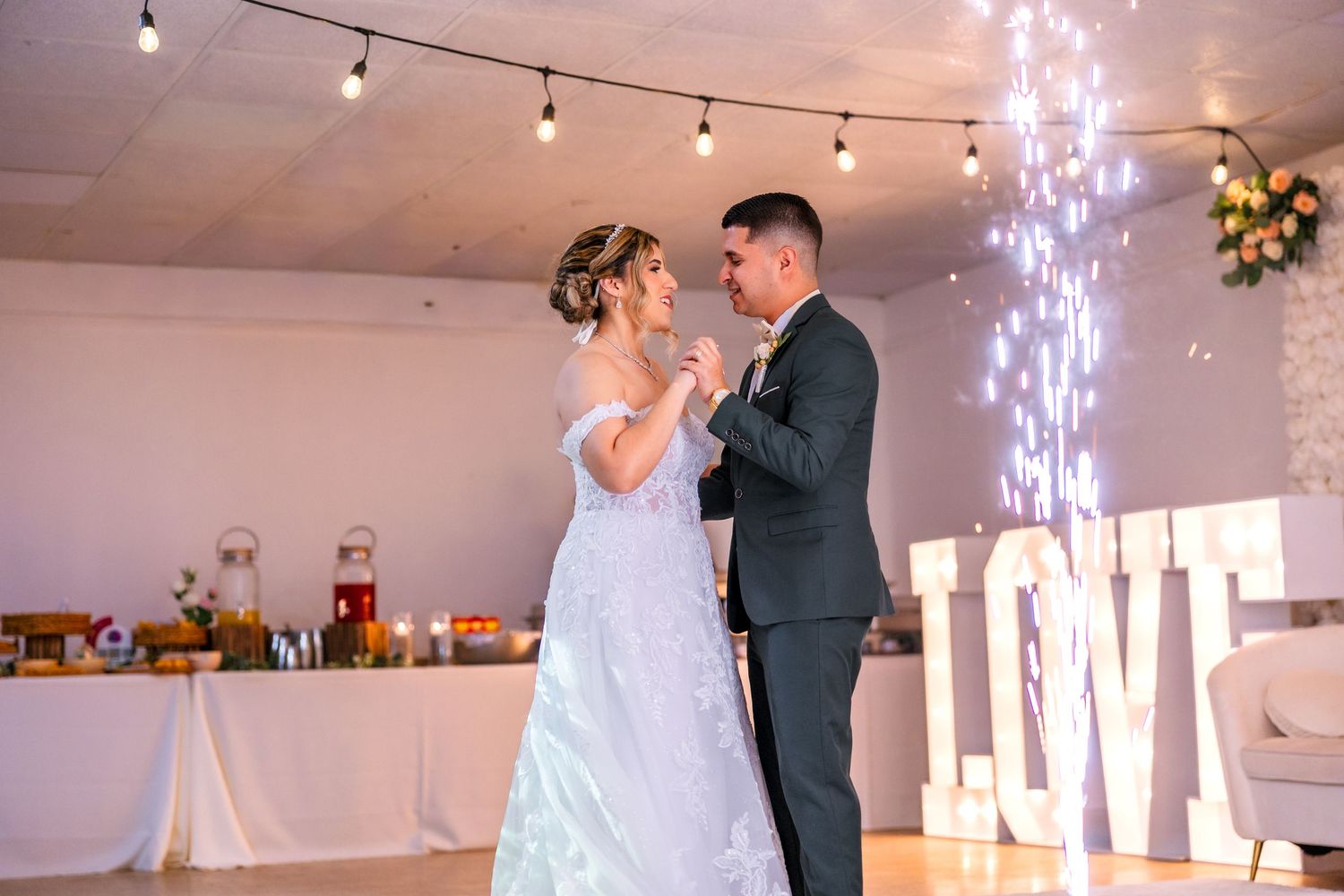 Bride and groom share first dance near illuminated LOVE letters with sparkling fountain at elegant wedding reception.