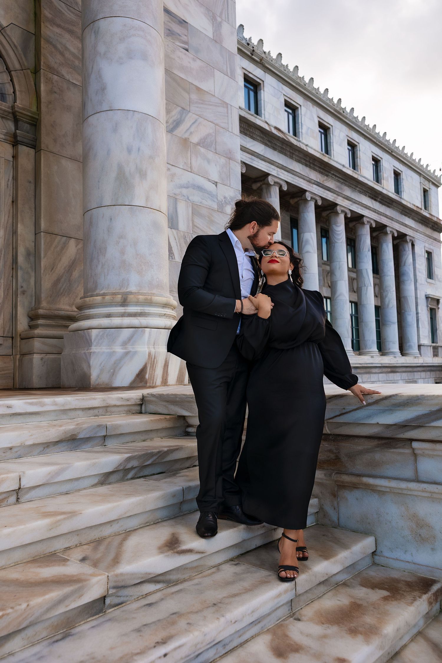 Couple in formal attire sharing a romantic moment on marble steps of a neoclassical building with columns.