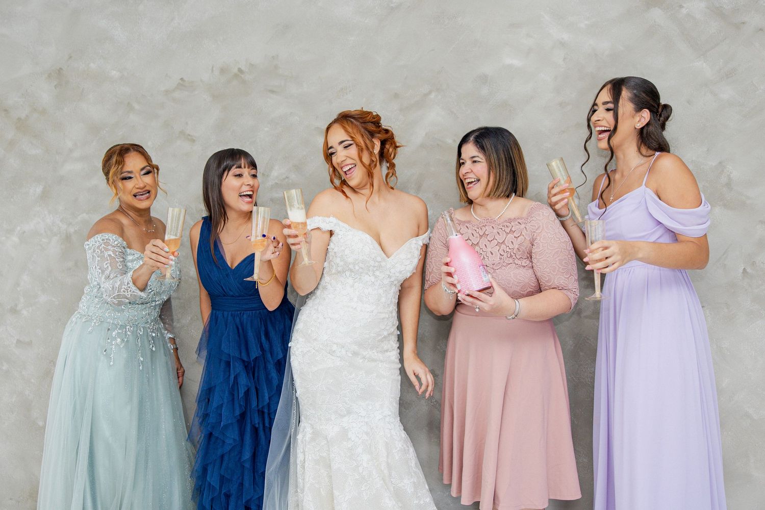A group of women in elegant formal dresses laugh and celebrate together against a textured wall.