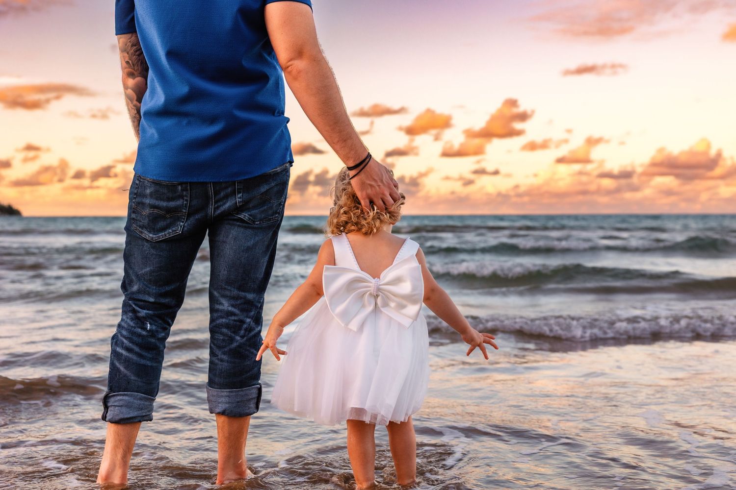Family walking along beach shoreline at sunset with waves crashing and golden sky in background.