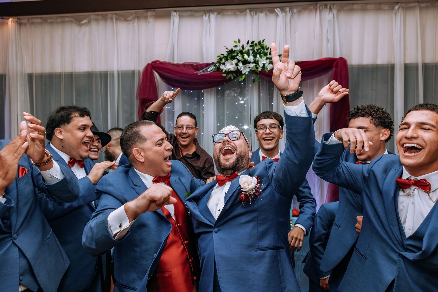 Joyful groom in blue suit celebrates with groomsmen in matching blue suits and red bow ties at wedding reception.