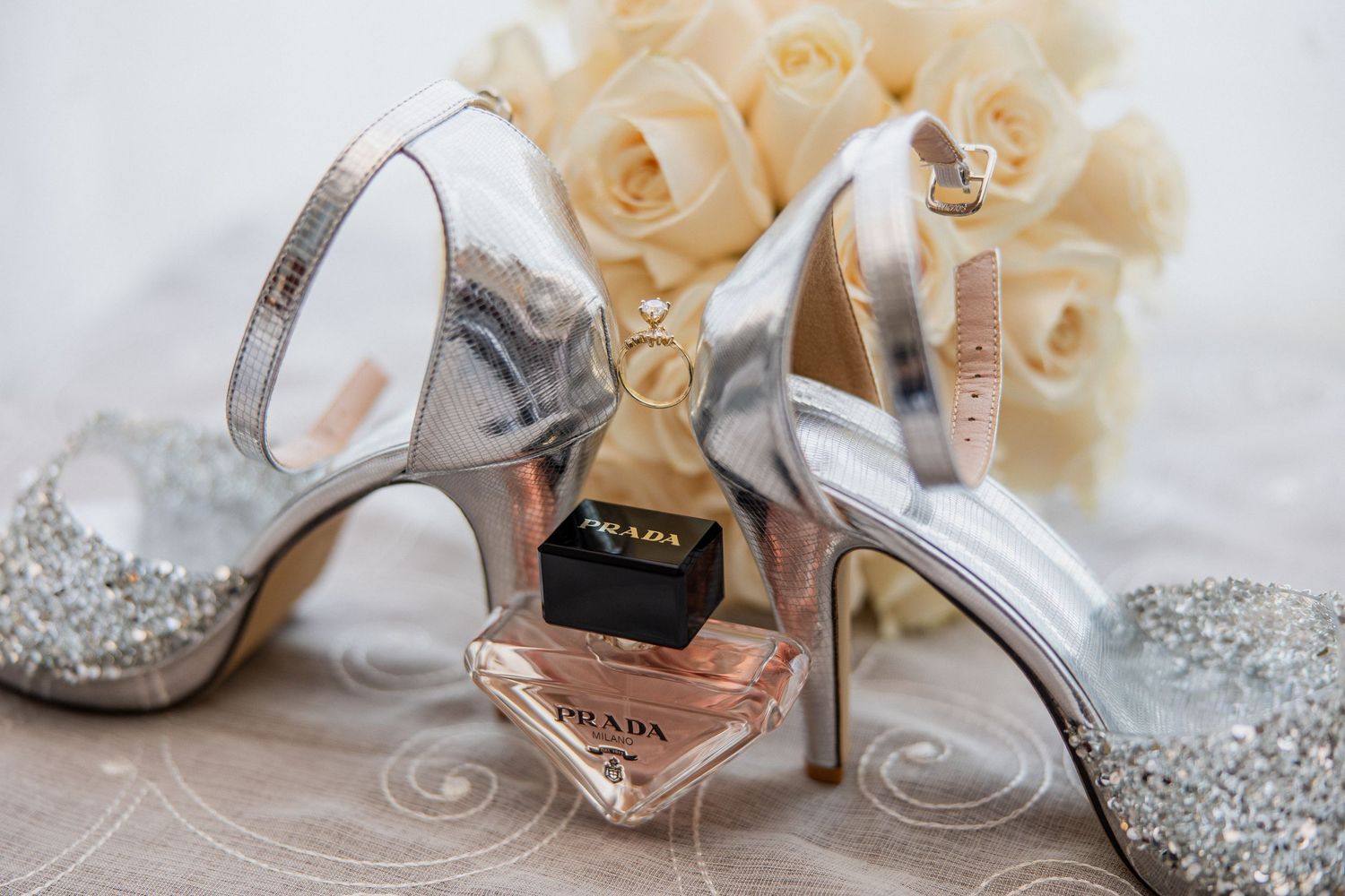 Silver glitter heels with Prada perfume bottle and ring box, surrounded by cream roses for a bridal flat lay.