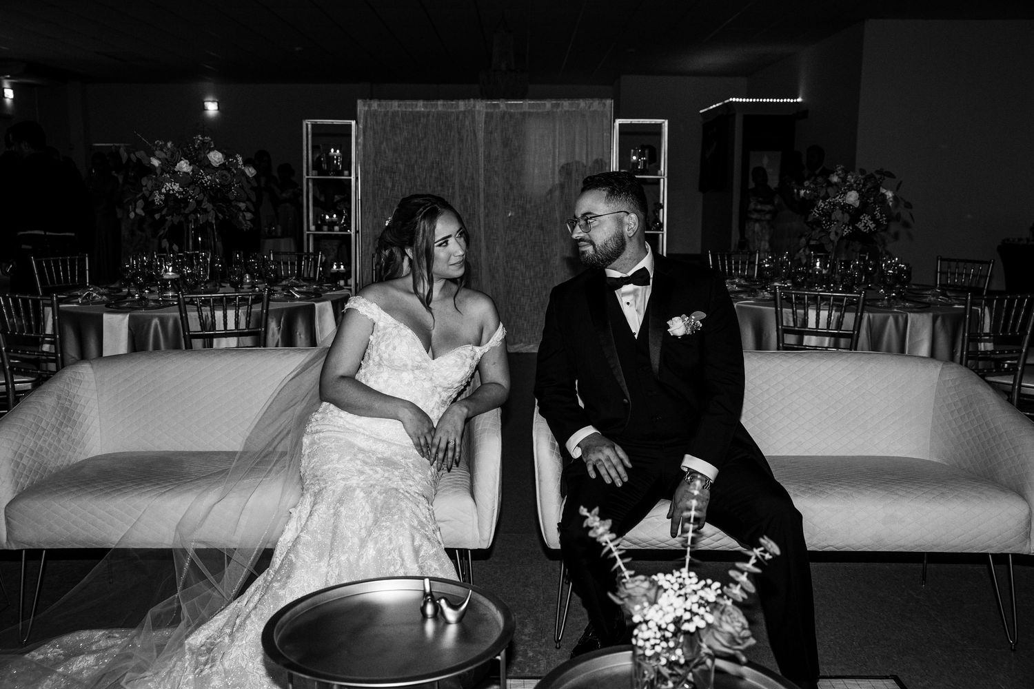 Black and white photo of bride and groom sitting on white sofa, sharing an intimate moment at their wedding reception.