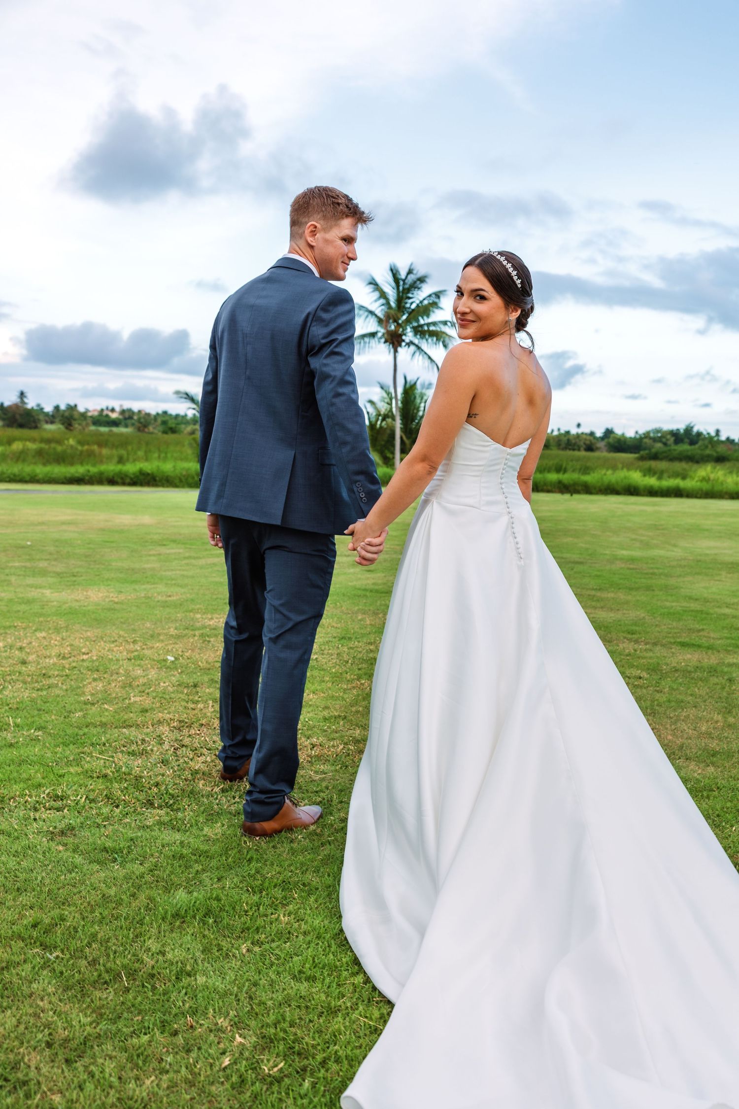 Bride in white gown looks back smiling while holding groom's hand on lush green lawn with tropical palms.