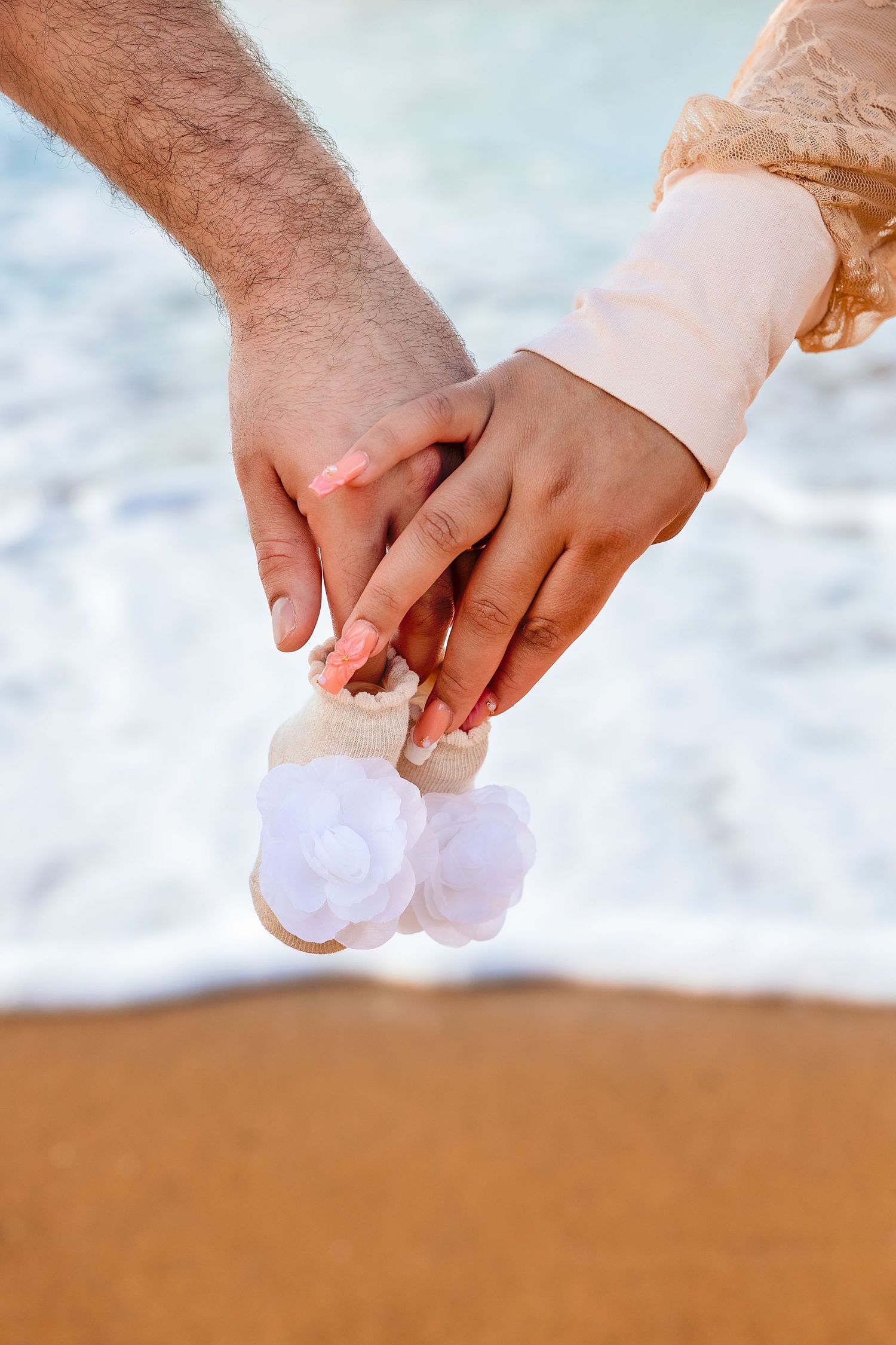 Couple holding hands together on sandy beach, holding tiny white flower baby shoes near ocean waves.