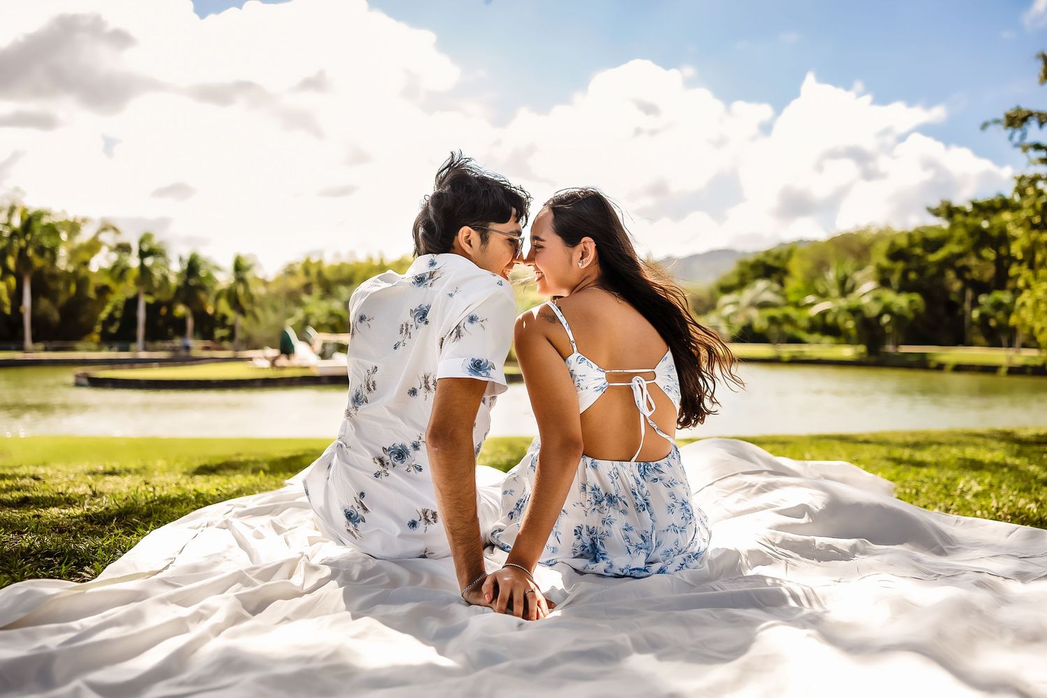 Couple sharing a romantic moment on a white blanket in a lush green park by a lake on a sunny day.