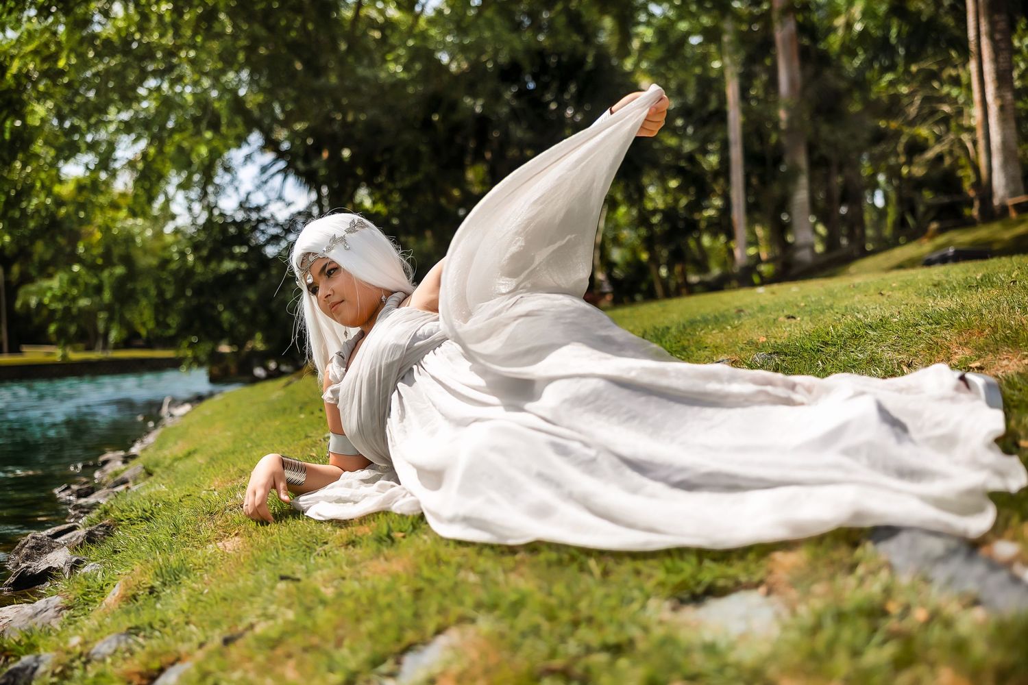 Woman in white hijab and flowing dress poses gracefully on green grass beside a tranquil pond in a lush garden.