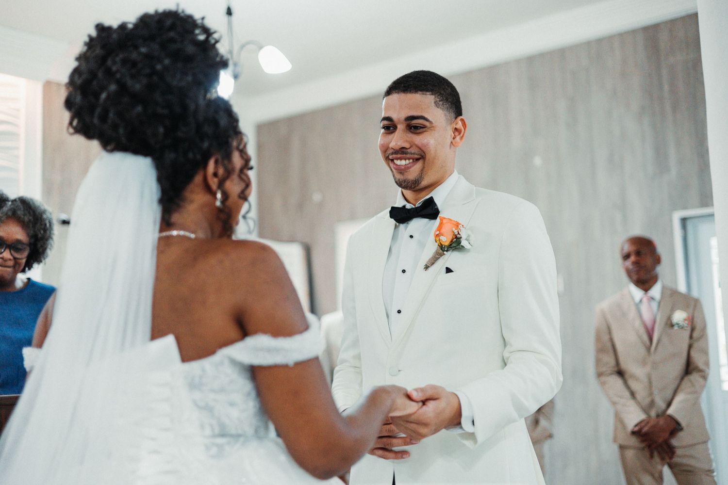 Groom in white tuxedo smiling at bride in wedding dress during indoor ceremony with guests in background.