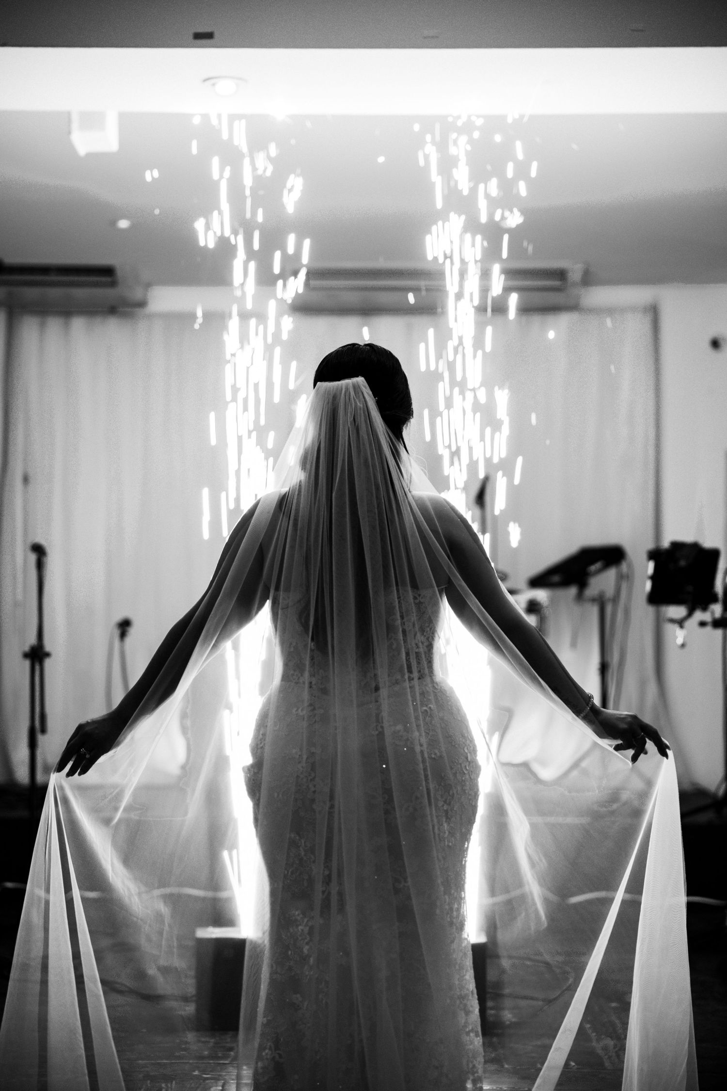 Black and white photo of a bride holding her veil wide open, backlit by sparkling lights at a wedding reception.