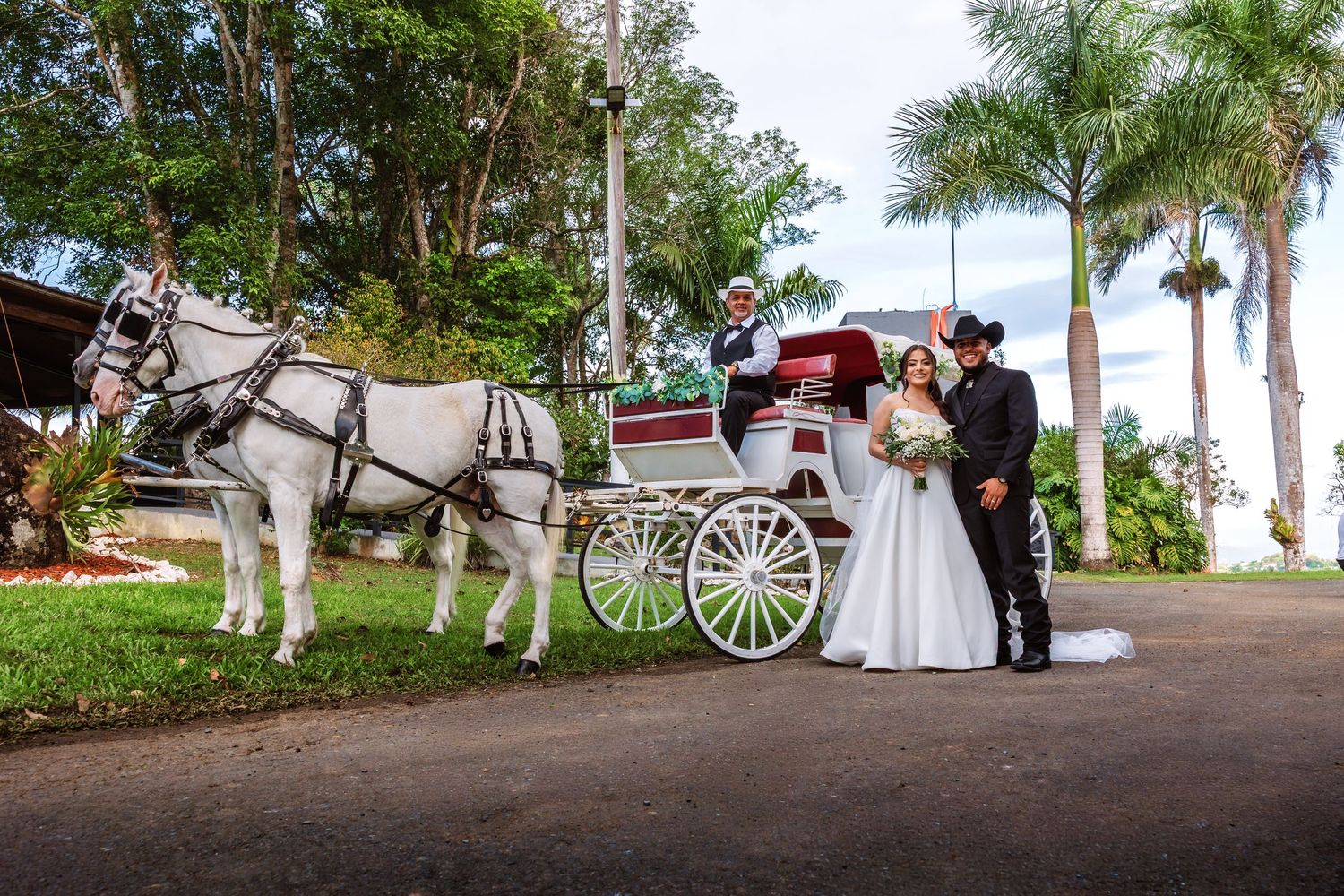Bride and groom pose beside a white horse-drawn carriage with a coachman at a tropical outdoor wedding venue.