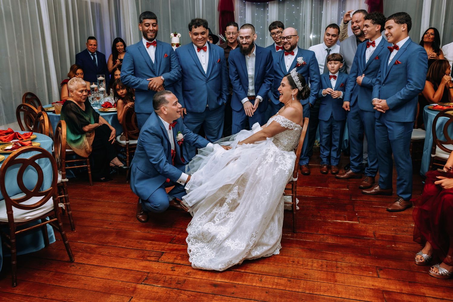 Groom kneels before bride in wedding gown during reception garter ceremony, surrounded by groomsmen in blue suits.