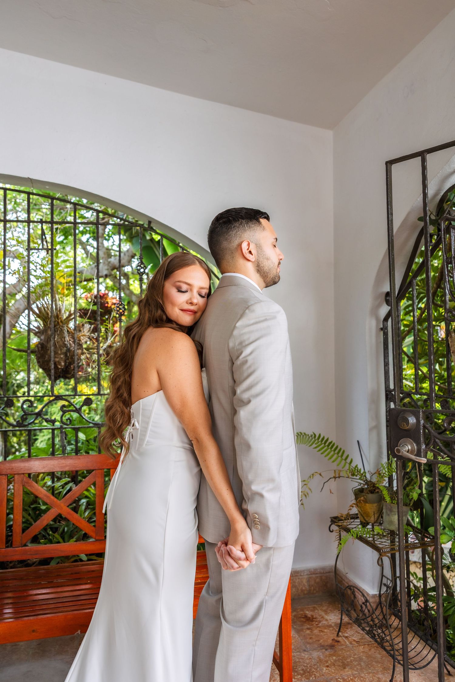 Bride and groom holding hands back-to-back in elegant wedding attire near an arched garden gate.