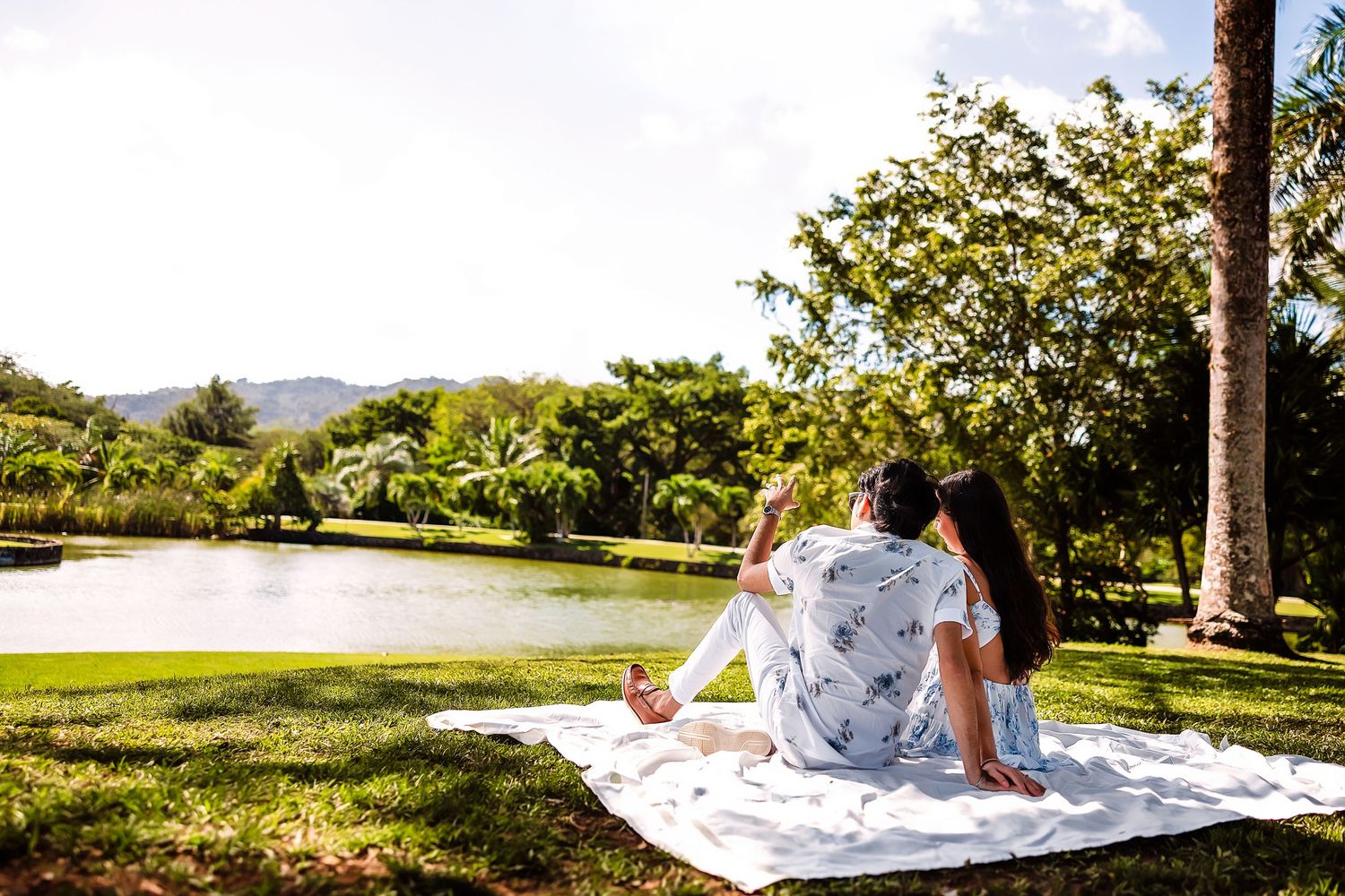 Couple sitting on white blanket enjoying scenic lake and mountain views in lush tropical park.