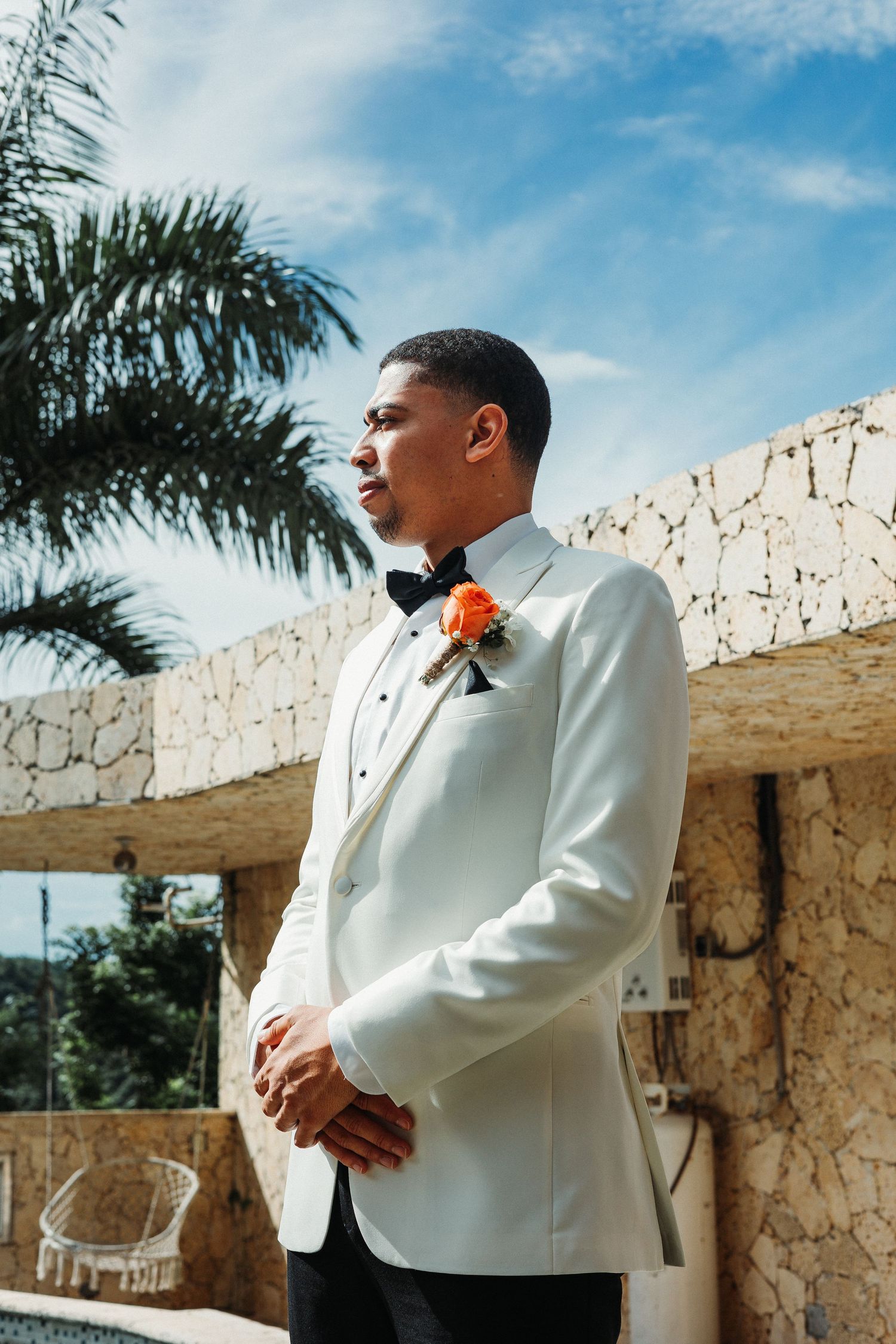 Groom in white tuxedo jacket with orange boutonniere stands outdoors near palm trees on a sunny wedding day.
