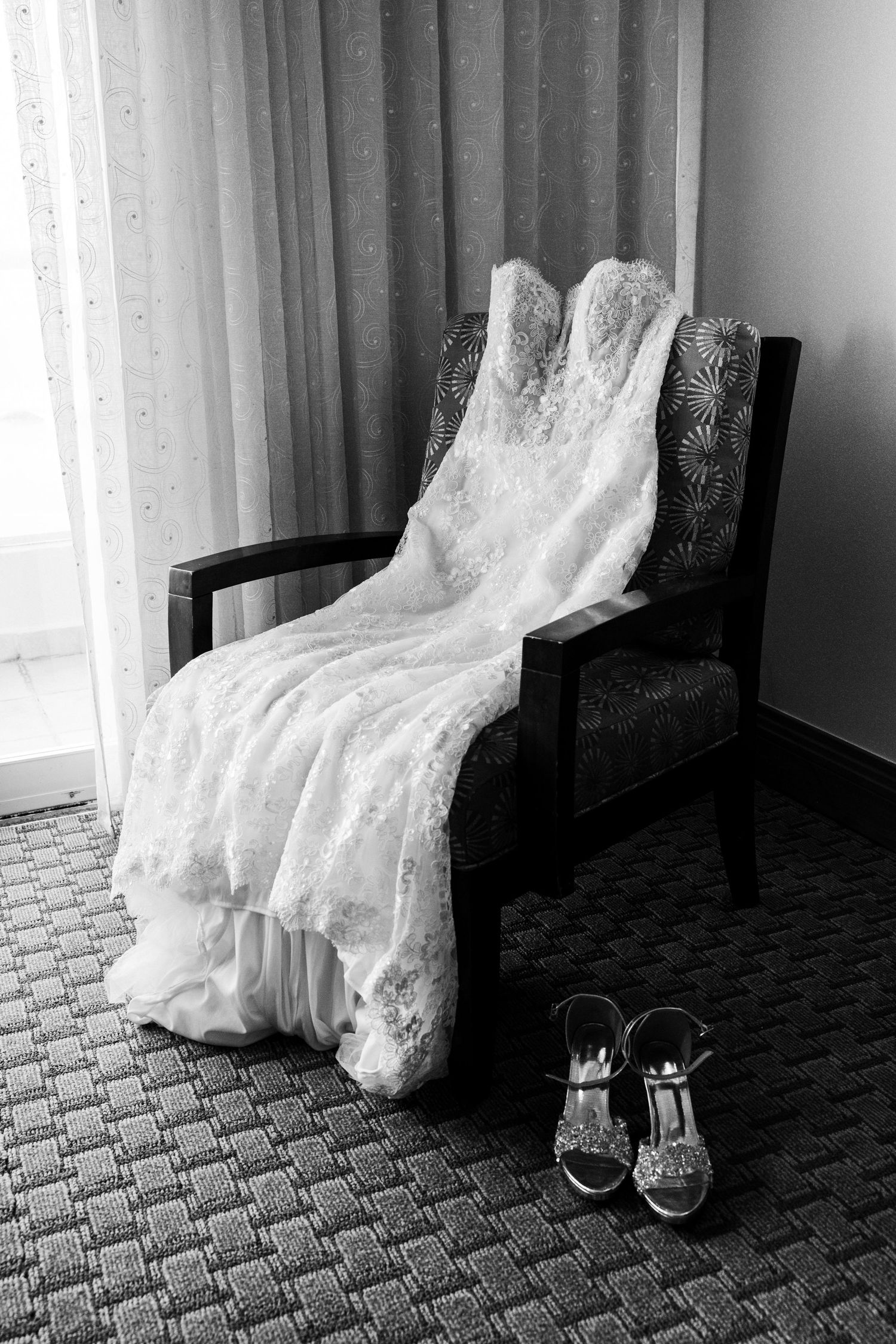 Black and white photo of a lace wedding dress draped over a chair with bridal heels on the floor beside it.