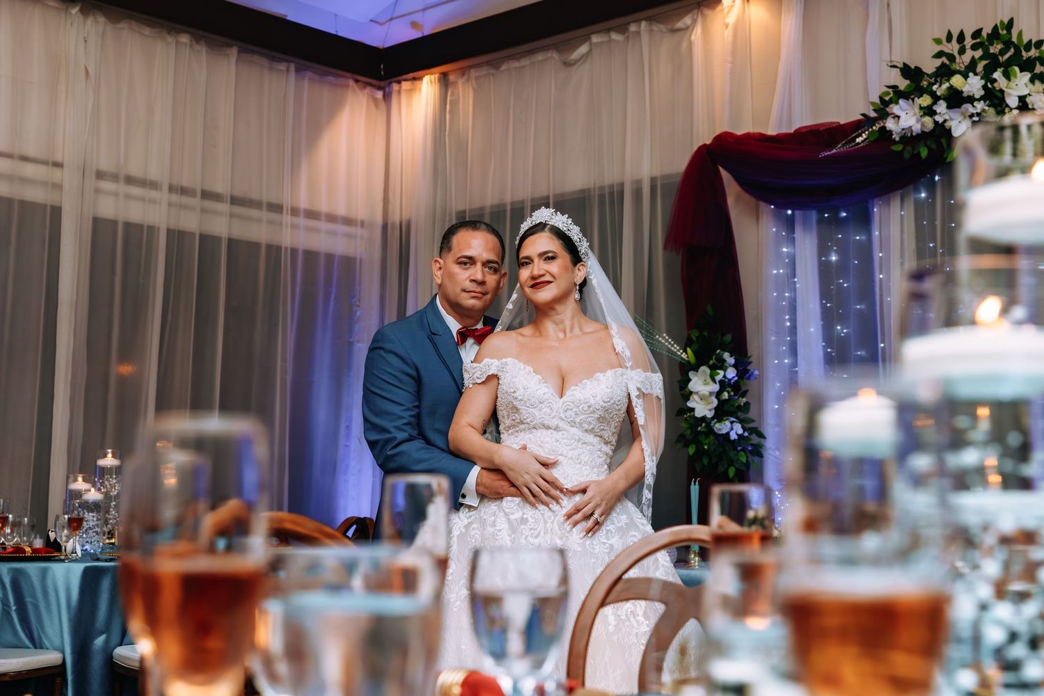 Bride and groom posing together at elegant wedding reception with draped curtains and candlelit tables in background.