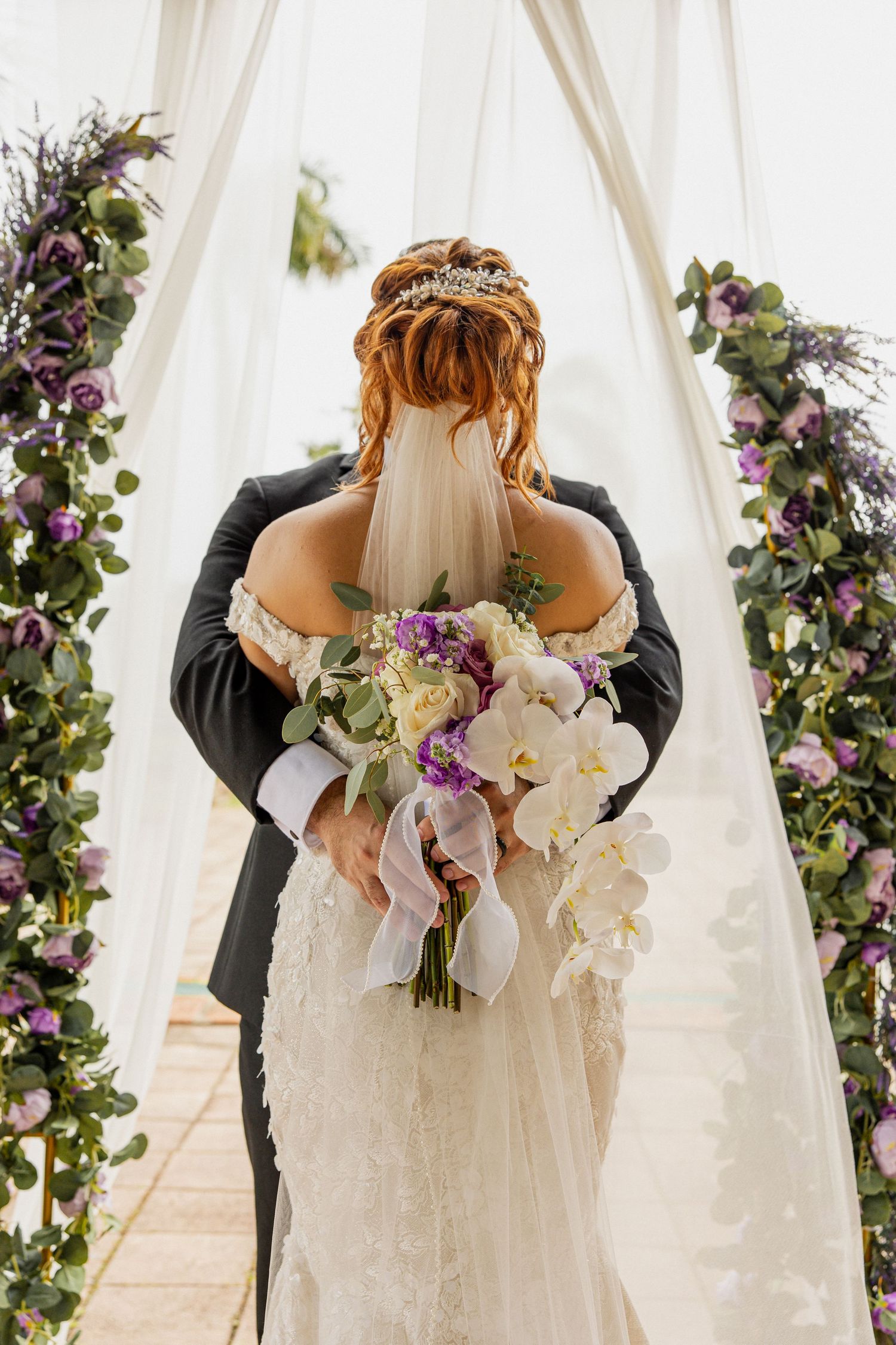 Bride and groom embracing at floral wedding altar with purple flowers, greenery, and white draping, bride holding bouquet.