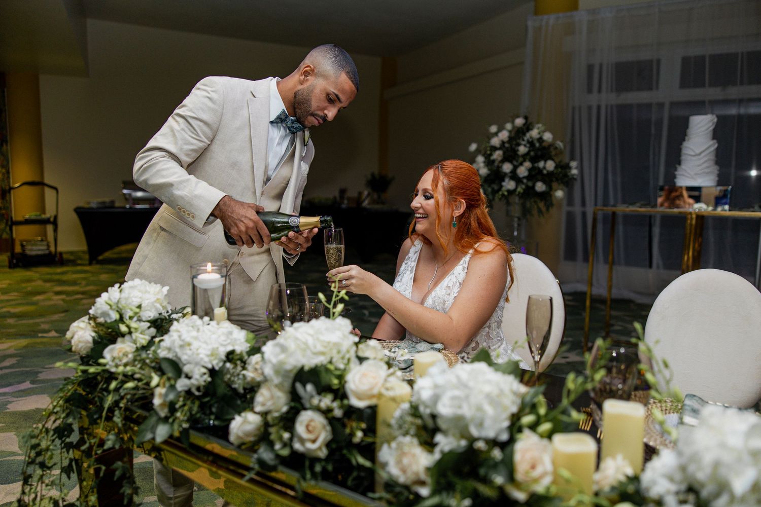 Groom in white suit pouring drinks for smiling bride at elegant wedding reception table with white floral arrangements.