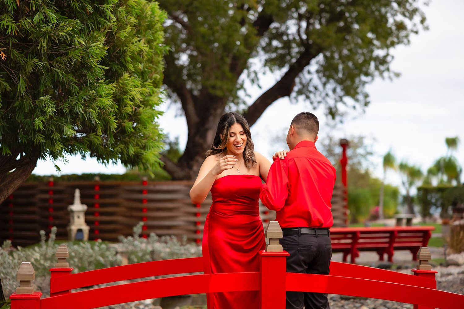 A couple in matching red outfits stands on a decorative red bridge in a Japanese garden setting.