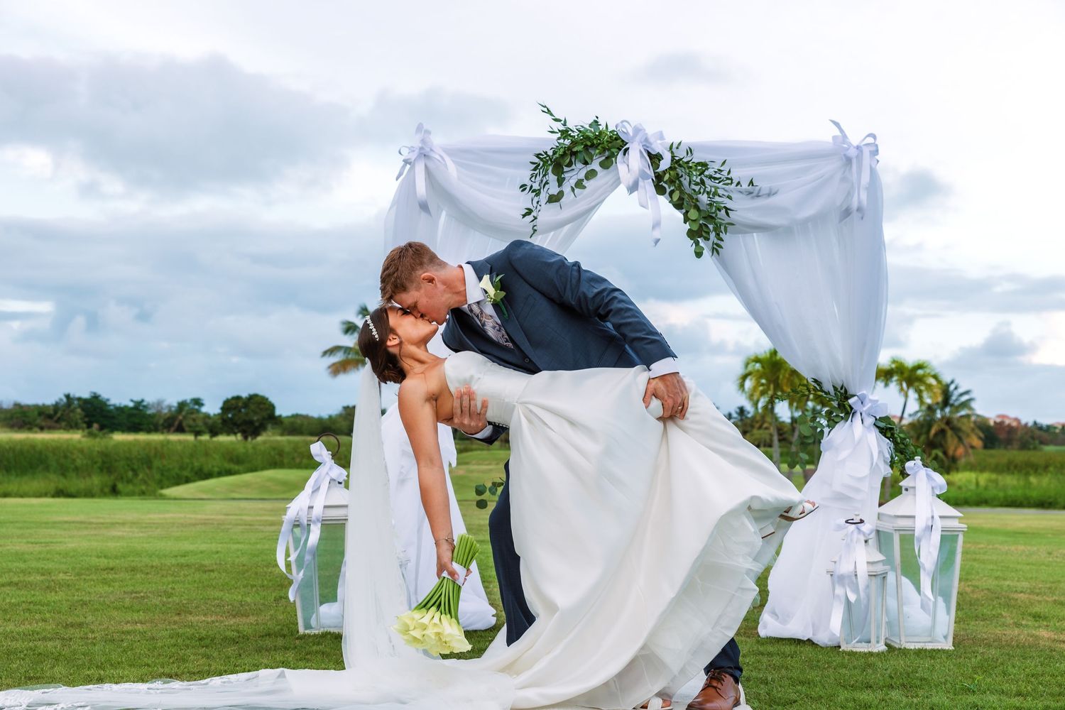 Groom dips bride in romantic kiss under white floral arch at outdoor wedding ceremony on lush green golf course.