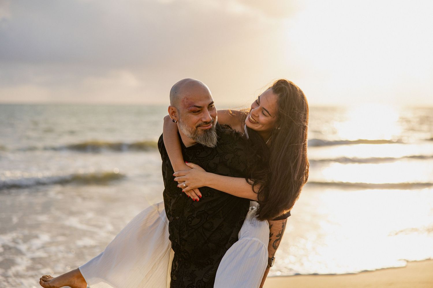 Couple embracing on a beach at sunset, man carrying woman as waves crash behind them in golden light.
