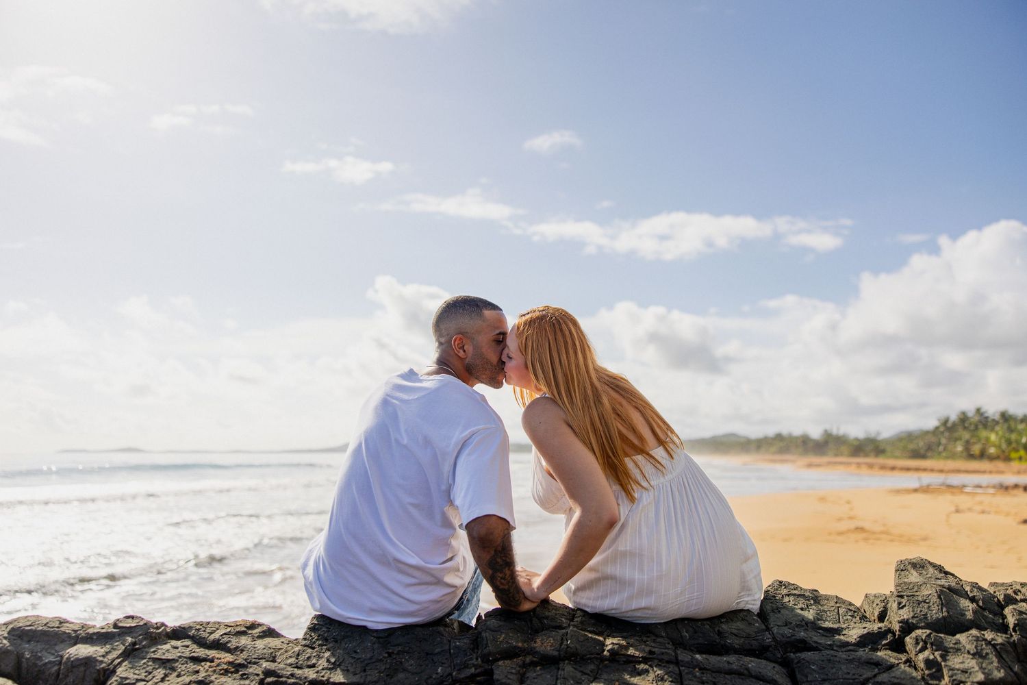 Couple sharing a romantic kiss on rocky shore with sandy beach and ocean waves in the background.
