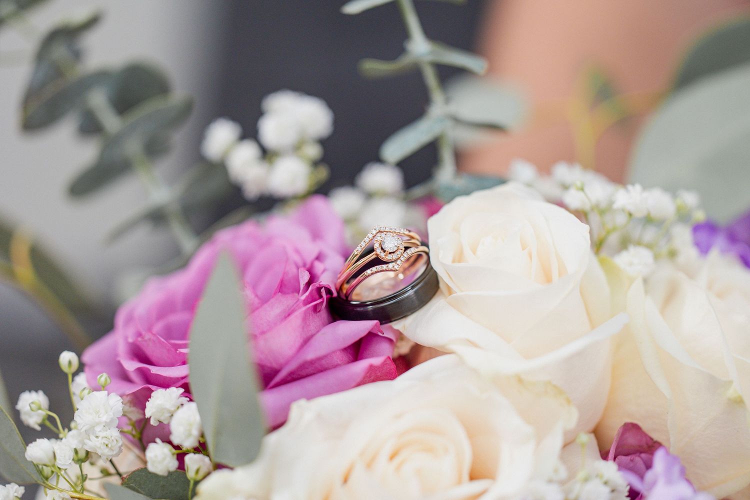 Two gold wedding rings rest on a bridal bouquet of white and purple roses with eucalyptus greenery.