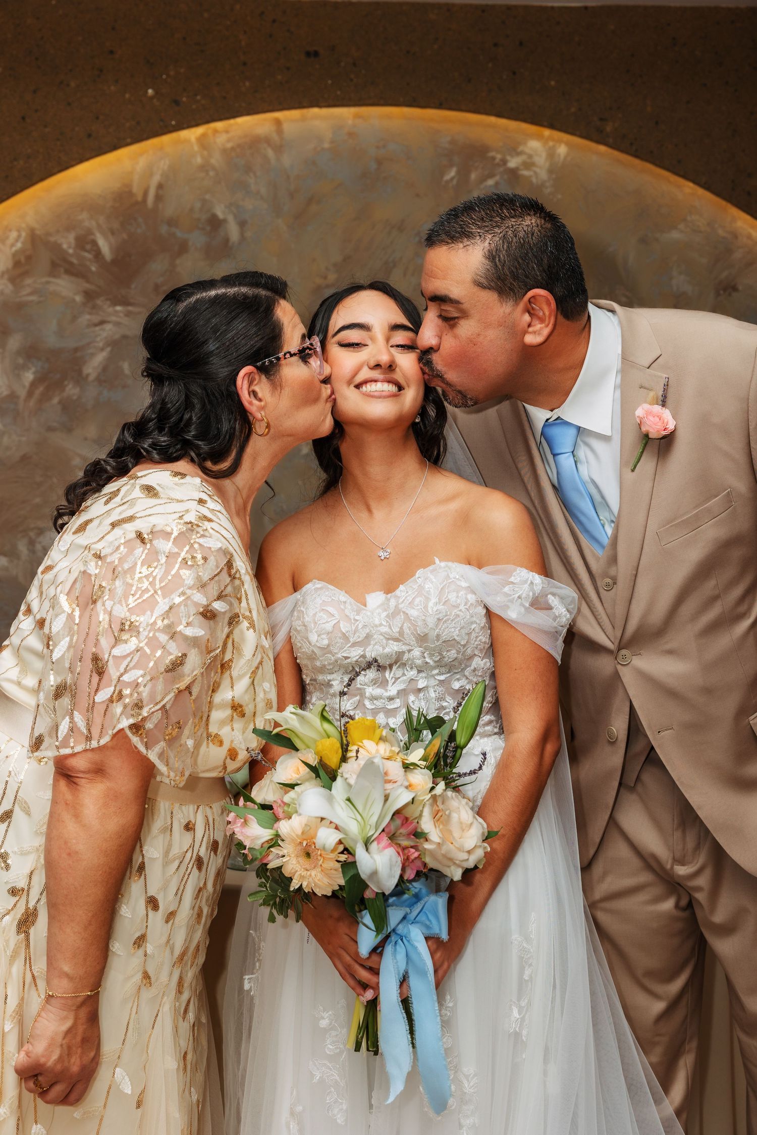 Bride smiles as parents kiss her cheeks on wedding day, holding a pastel bouquet in an elegant venue.