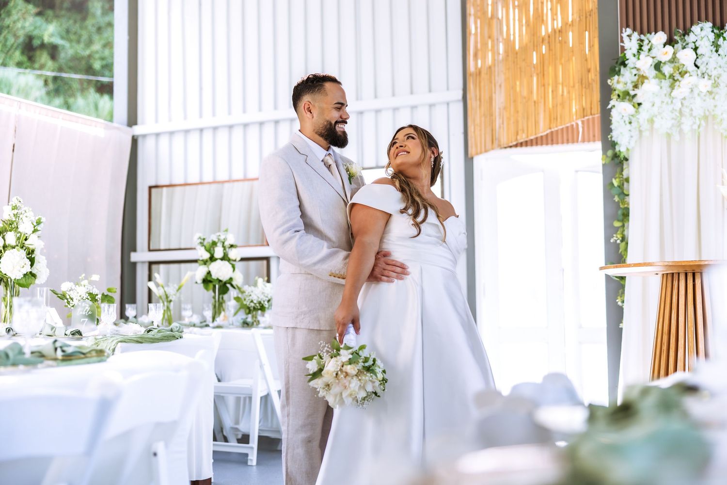 A happy couple in wedding attire embraces at an elegant white-decorated venue, the bride holding a bouquet.