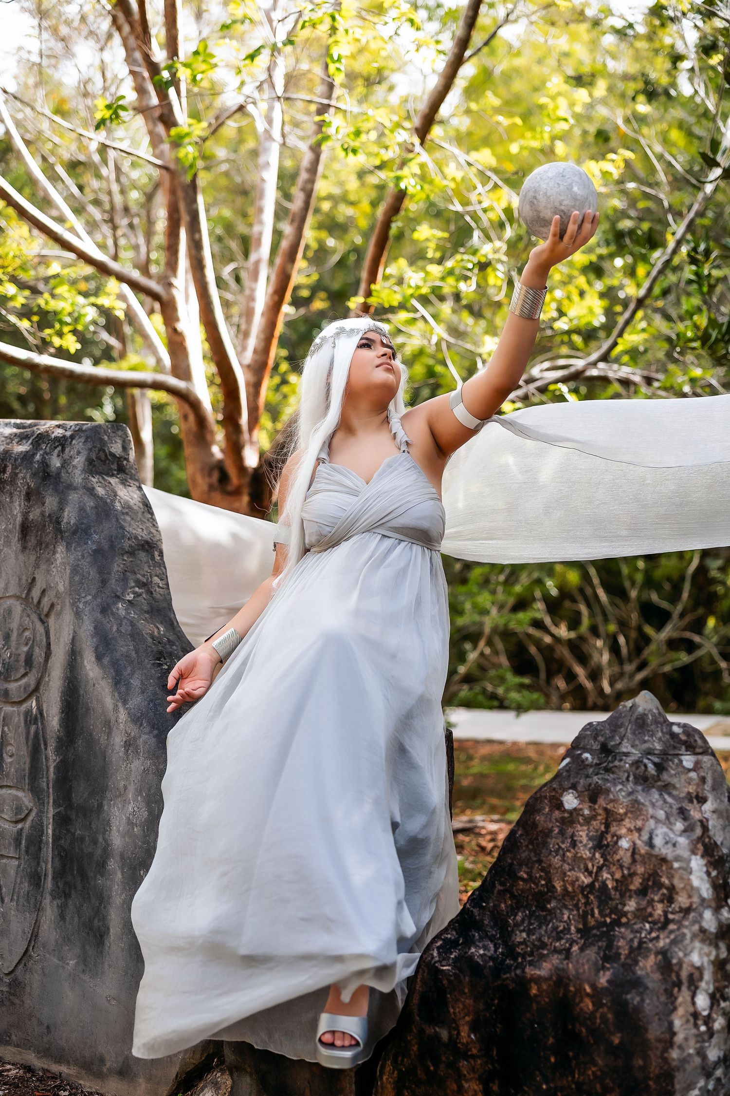 Woman in white dress holding crystal ball among ancient stones in lush forest setting.